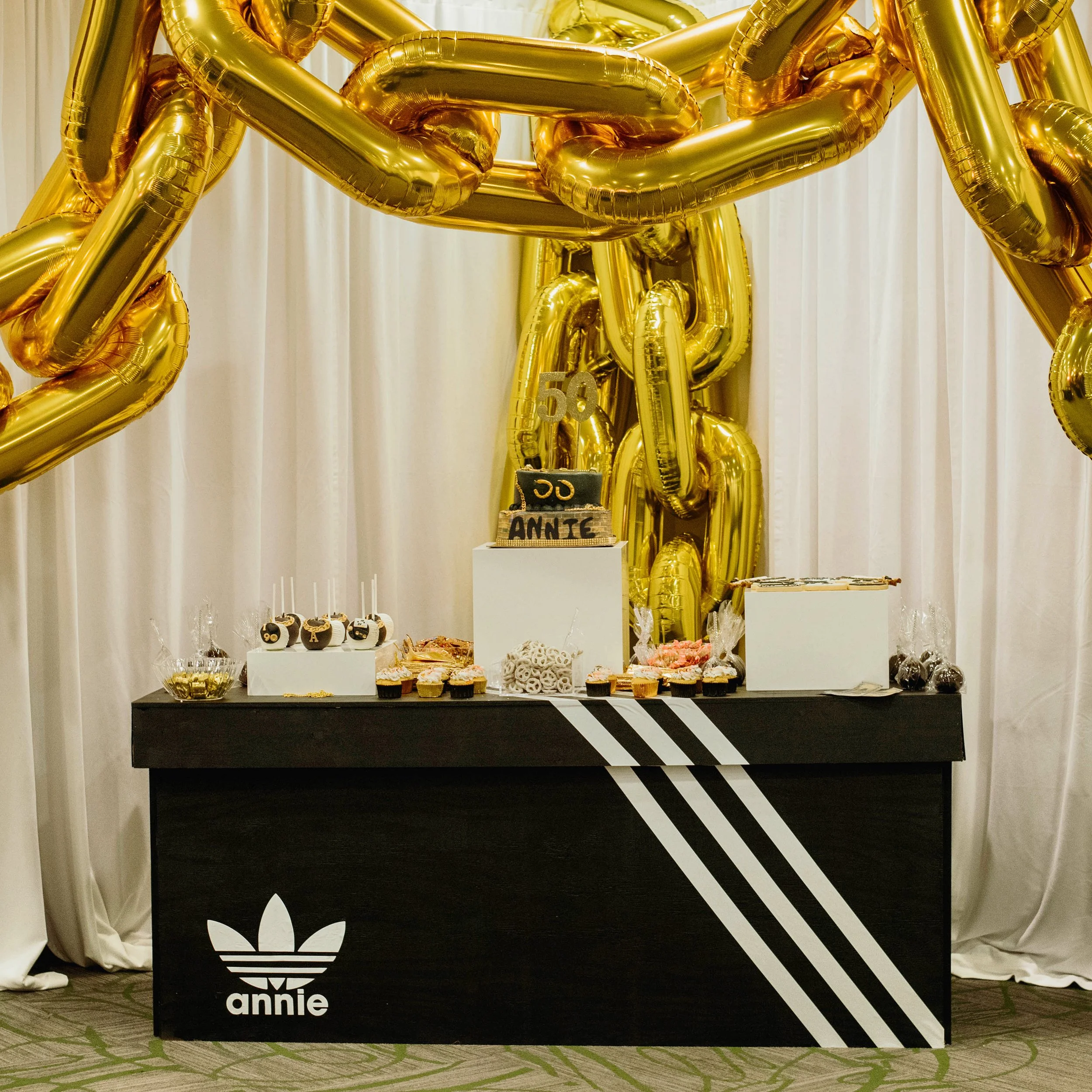 Dessert table decorated with large gold balloons, a birthday cake, cupcakes, and candies at a celebration event.