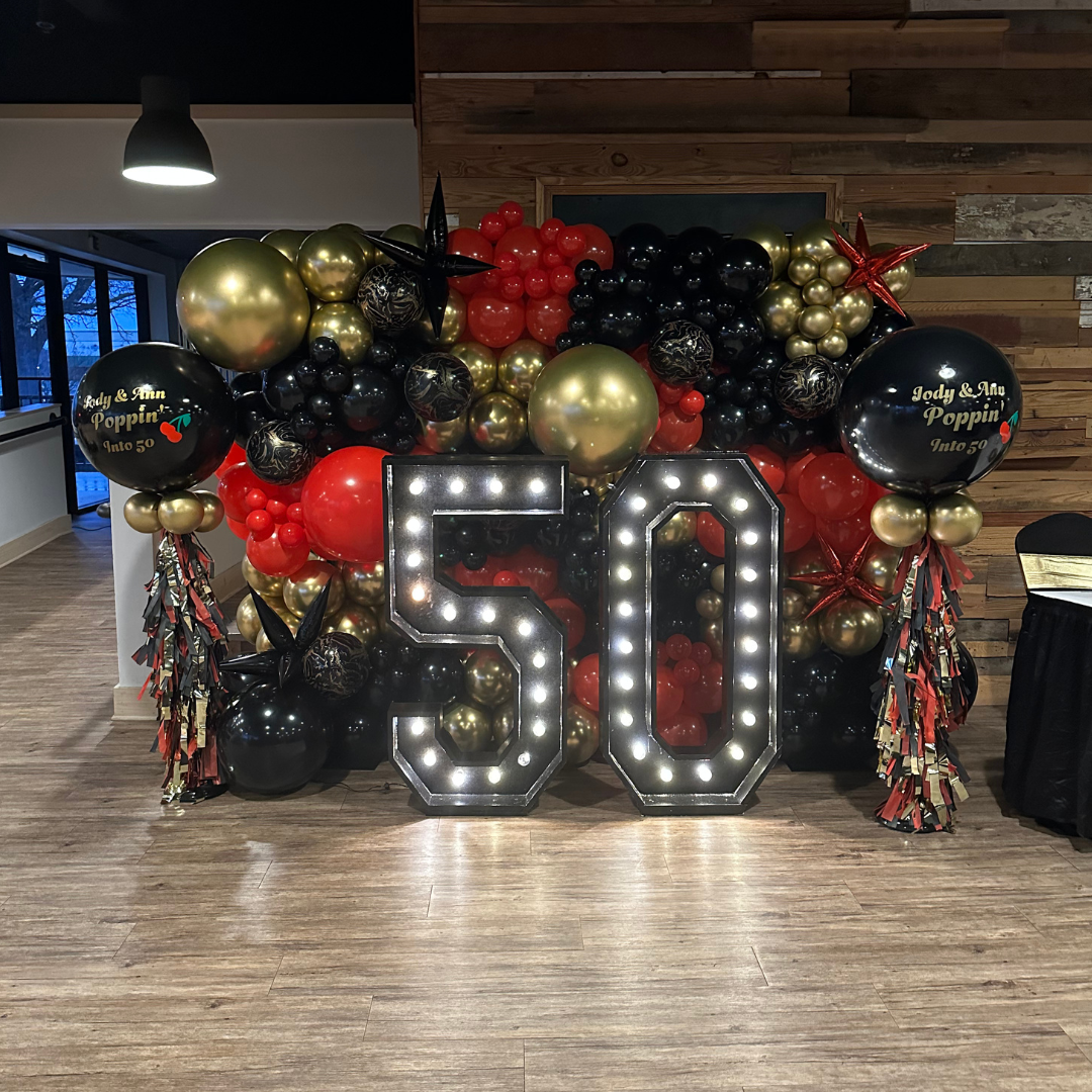 Decorative celebration display featuring a large illuminated '50' sign, black, gold, and red balloons, star-shaped balloons, and a balloon saying 'Jody & Ann Poppin' Into 50', set against a wooden wall backdrop.