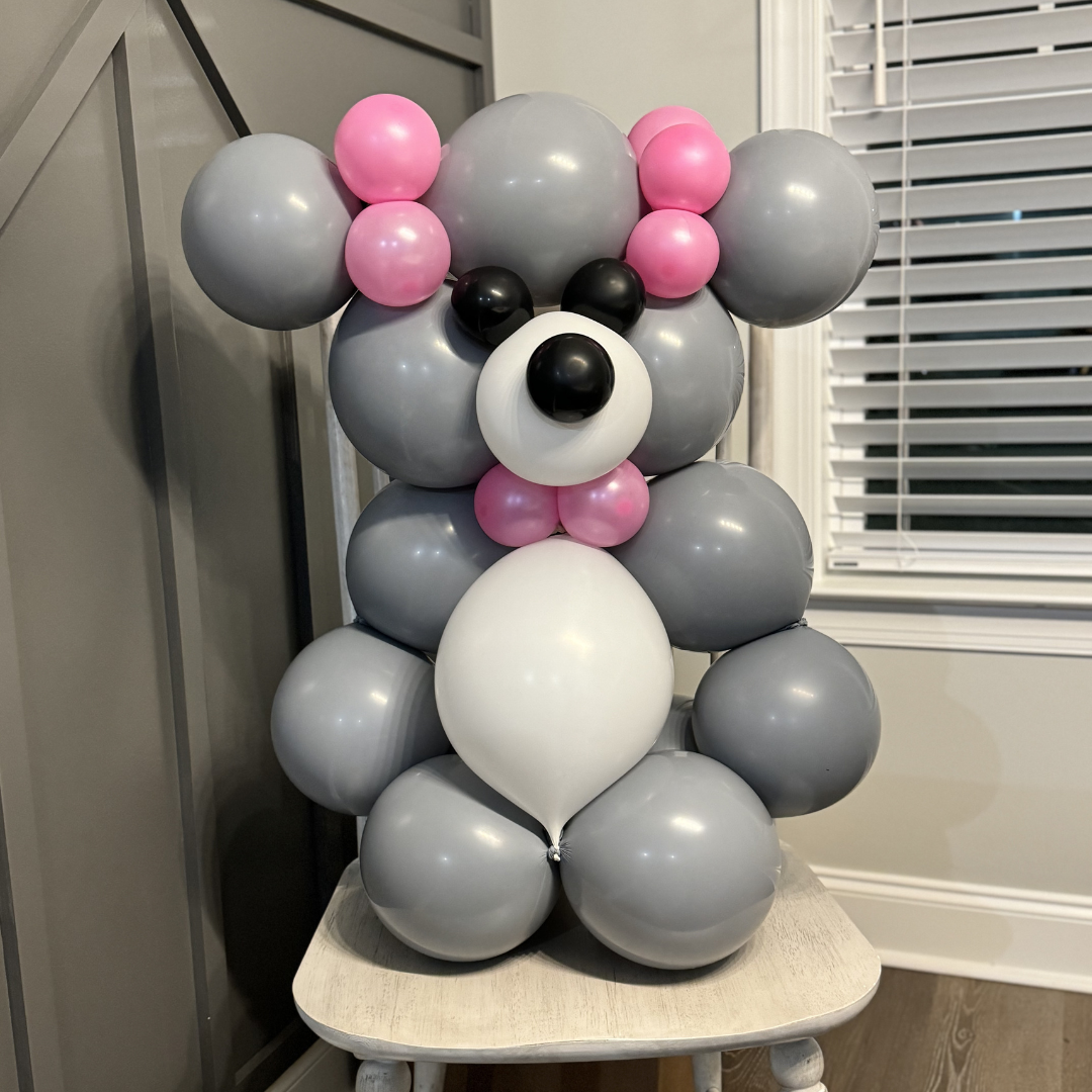 Balloon sculpture of a cute dog with pink bows, black eyes, nose, and ears, in gray, white, black, and pink colors.
