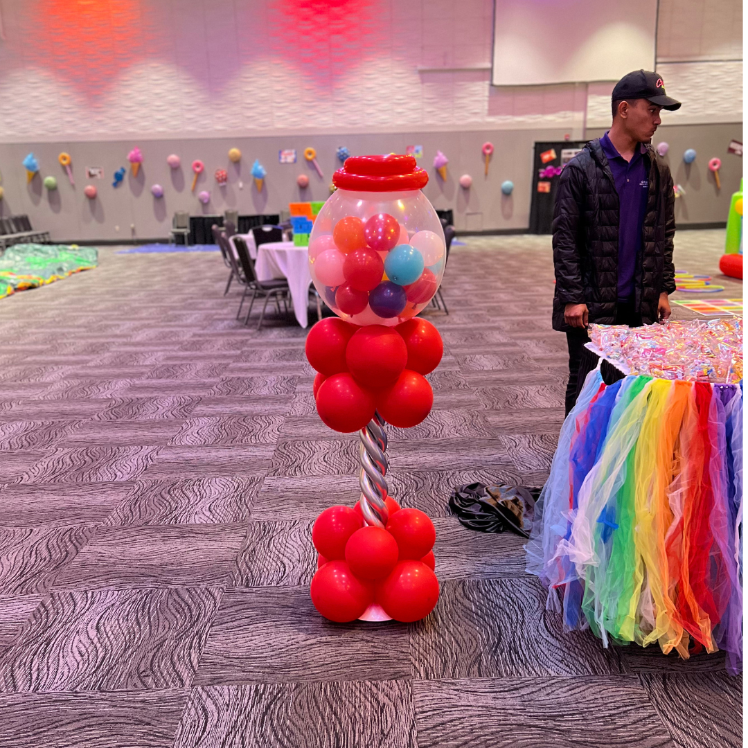 Colorful gumball machine decoration with red, pink, blue, and purple balls in a glass container, set on a base of red balloons, in an indoor party venue decorated with rainbow-colored tulle and background wall adorned with paper ice cream cones and c