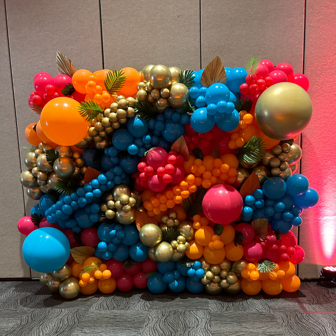 Colorful balloon display with various sized balloons in orange, blue, pink, red, and gold, decorated with fake leaves, set against a gray wall at an indoor event.