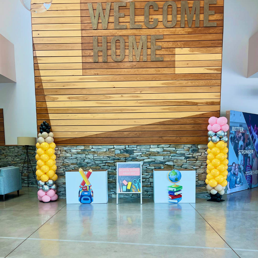 Decorative indoor welcome setup with balloon columns, school supplies, and a sign for school supply drop-off, against a wooden wall with 'Welcome Home' lettering.