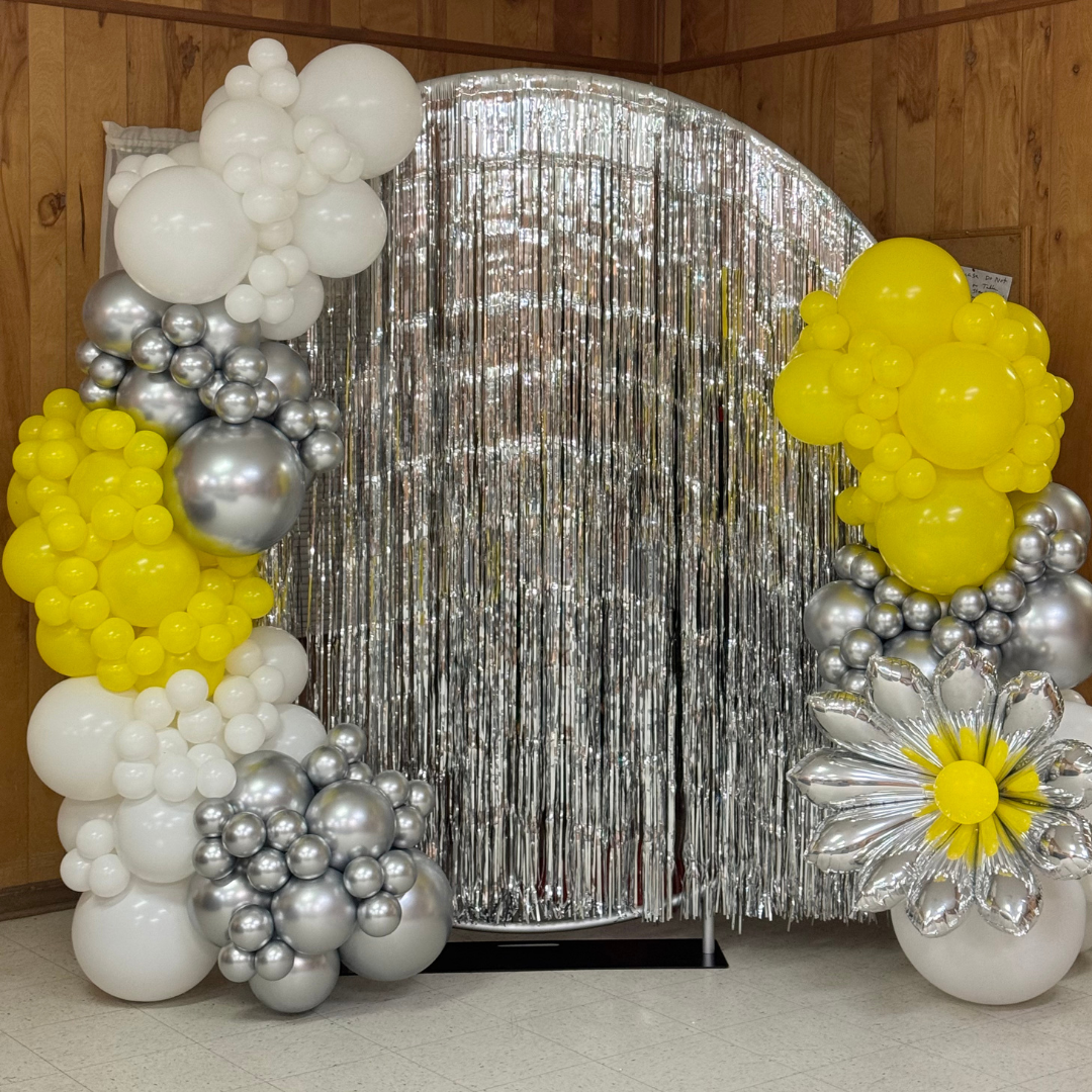 Decorative balloon arrangement with white, yellow, and silver balloons, silver fringe curtain backdrop, and a large silver flower balloon with yellow center.