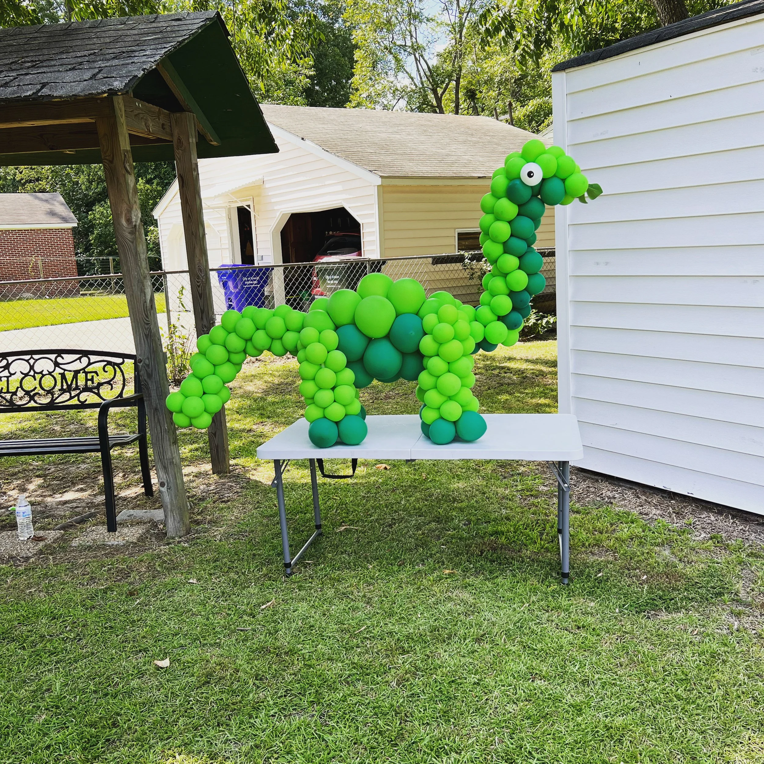 A dinosaur sculpture made of green and blue balloons standing on a white table outdoors in a backyard.