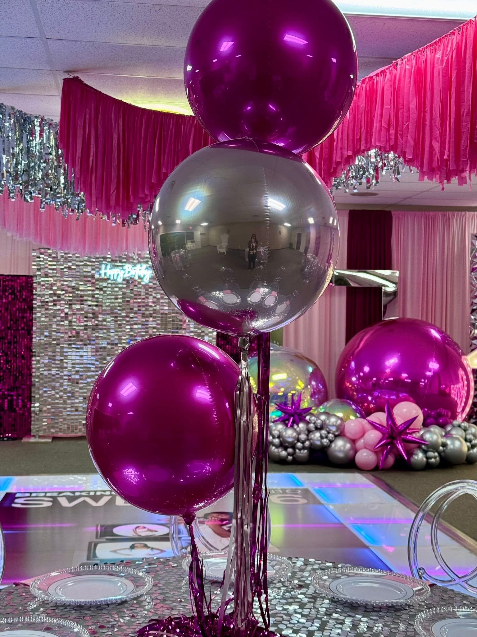 Pink, silver, and purple balloons decorated for a celebration, with pink curtains, silver tinsel, and a decorated backdrop in the background.