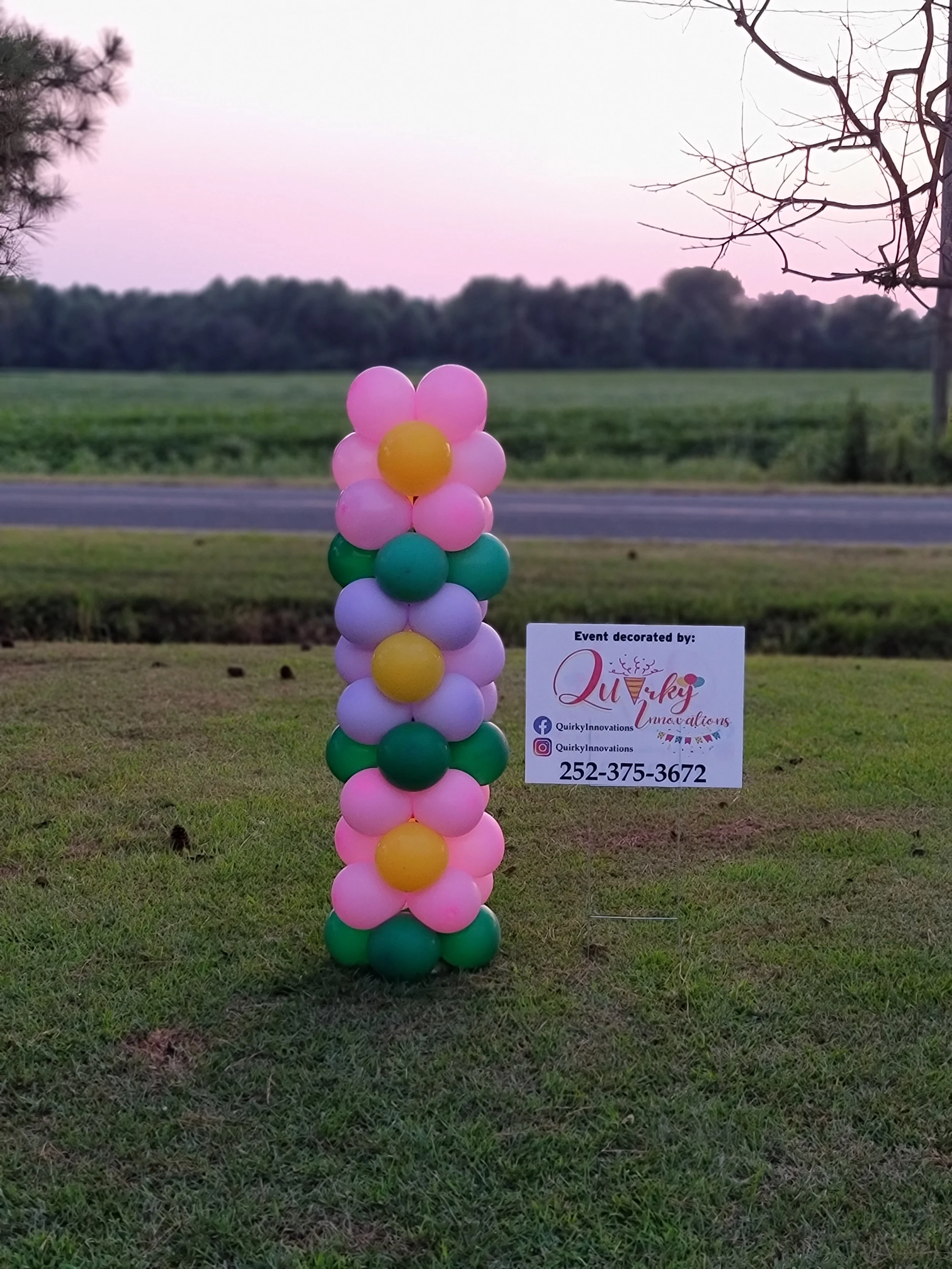 Colorful balloon arrangement of pink, green, and yellow balloons resembling a flower next to a sign on grass with trees and a road in the background.