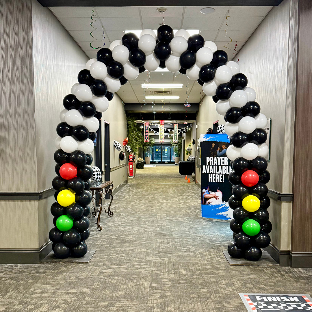Black, white, red, yellow, and green balloons arching over a hallway entrance decorated for an event, with a sign to the right that says 'Prayer Available Here!'