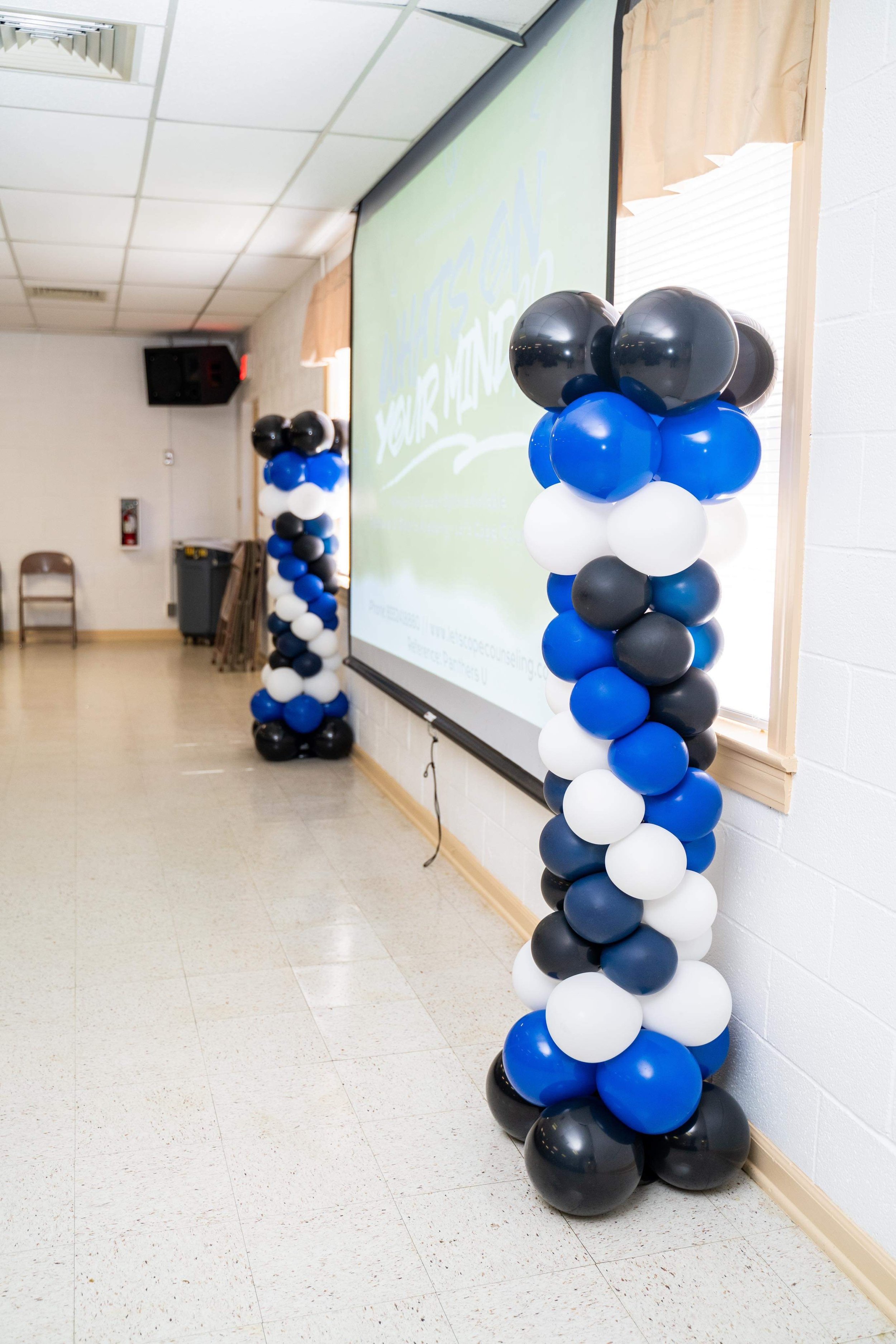Two columns of black, white, blue, and gray balloons border a presentation screen in a room with white walls and tile floor.
