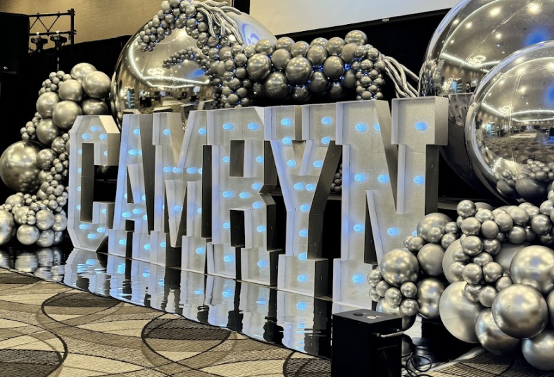 Decorative display with large illuminated letters spelling "CAMBYN" surrounded by silver and metallic balloons of various sizes.