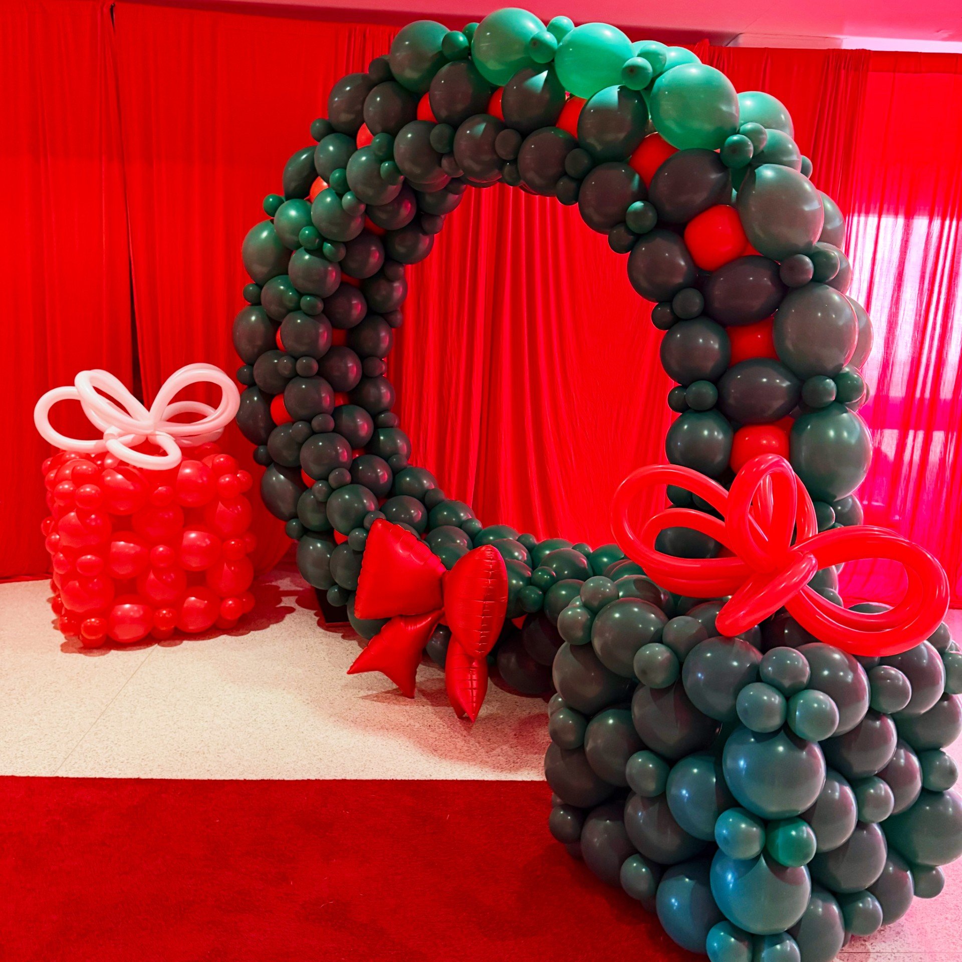 Decorative balloon arch and gift box made with balloons in red, green, and black colors, set against a red curtain background.