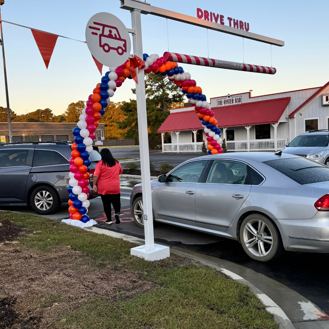 A woman entering a drive-through restaurant with an arch of colorful balloons and a sign that reads "Drive Thru." There is a restaurant building in the background with a sign that says "The Oyster Bar."