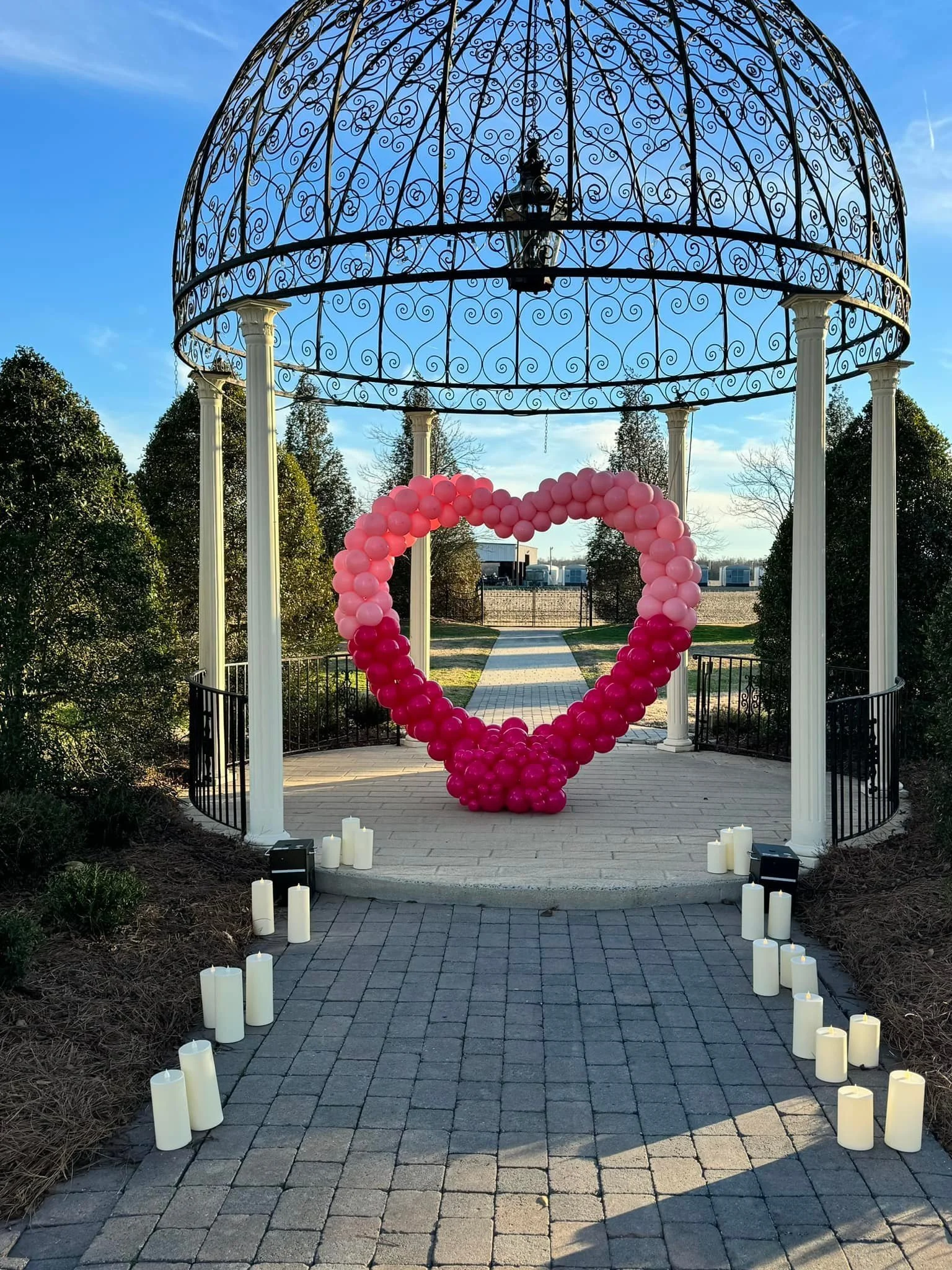 A gazebo with ornate ironwork and white columns, decorated with a heart-shaped balloon arrangement of pink and dark pink balloons, surrounded by candles on the ground, in an outdoor setting with a paved pathway and trees.