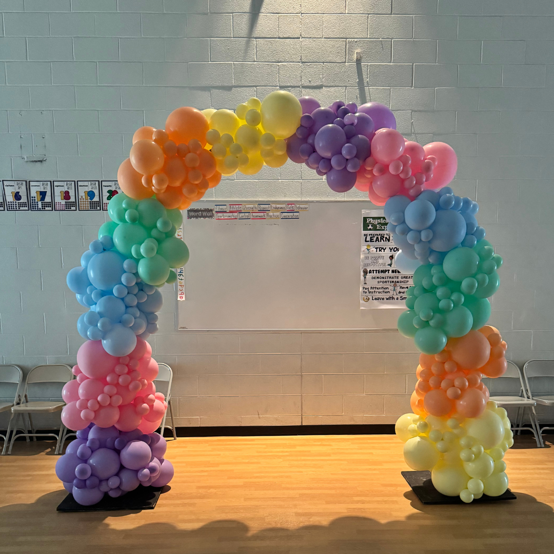 Colorful balloon arch with pastel colors including purple, pink, blue, green, yellow, and orange in a room with chairs and educational posters on the wall.