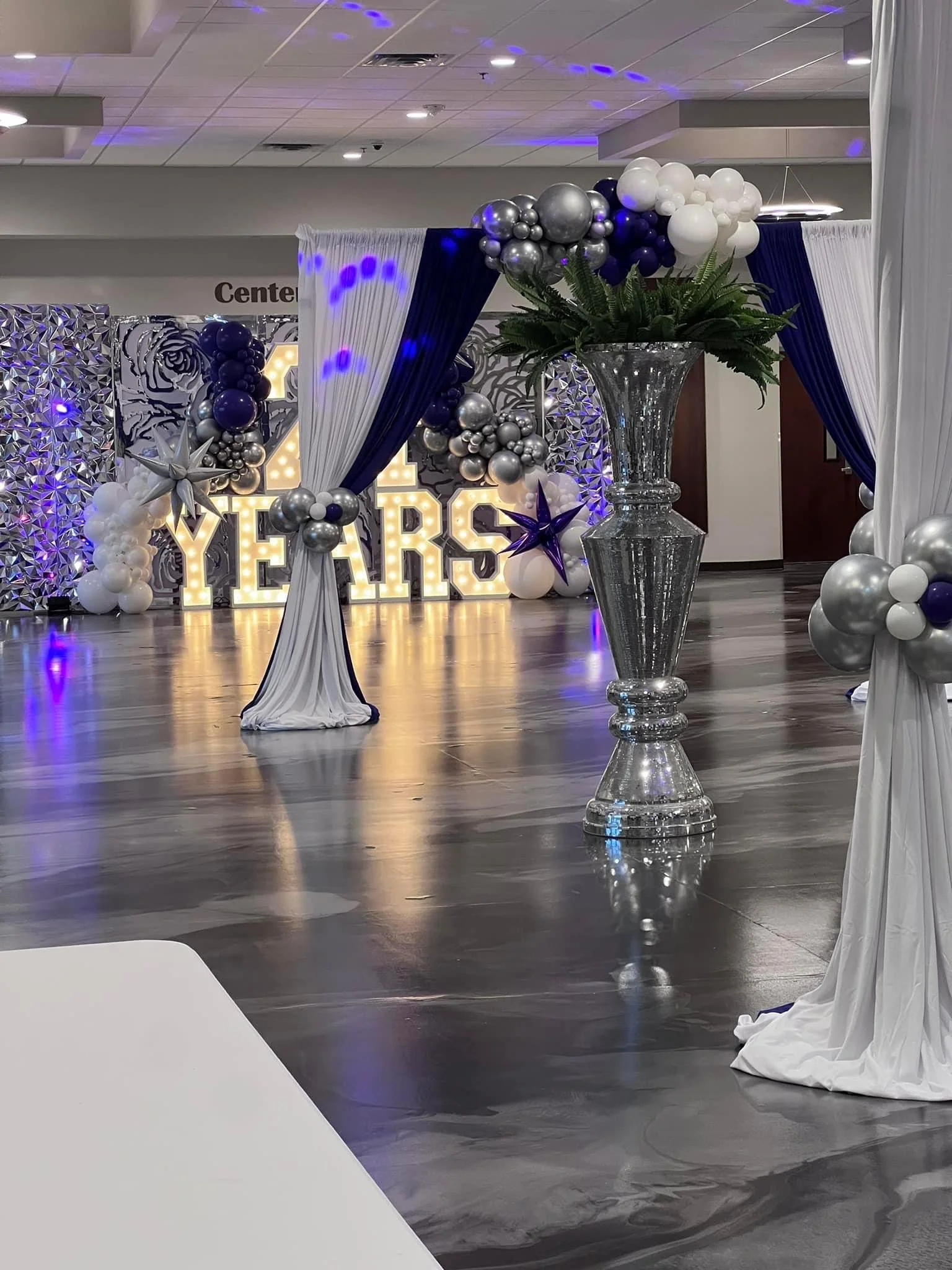 Decorative event setup featuring large illuminated sign spelling 'YERS,' surrounded by white, silver, and navy blue balloons, white and navy blue draped curtains, and large glass vases with floral arrangements. The setting appears to be for a celebra