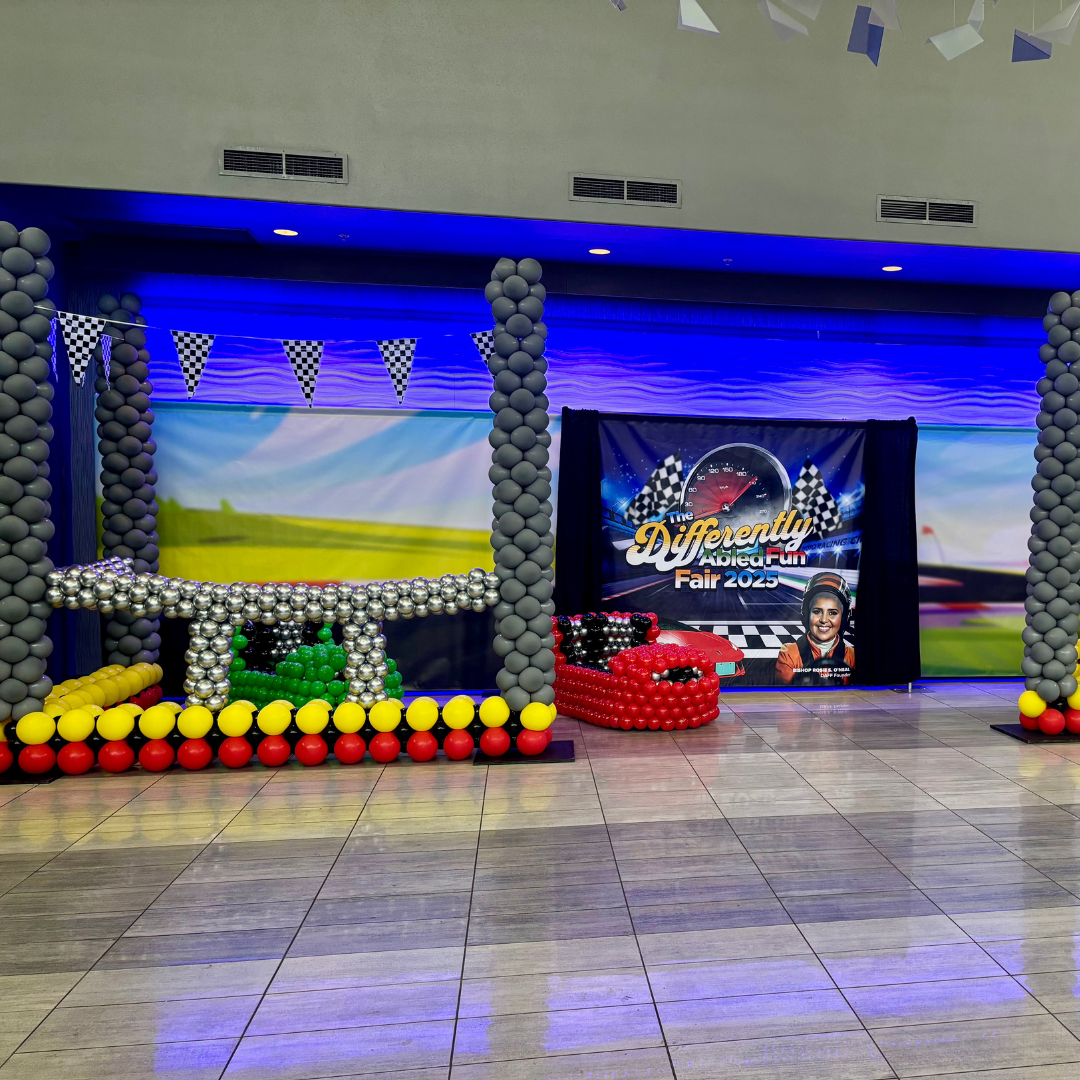 Decorative display with balloons and a racing-themed backdrop for the "Differently Abled Fun Fair 2025," featuring checkered flags, a speedometer graphic, and a smiling person.