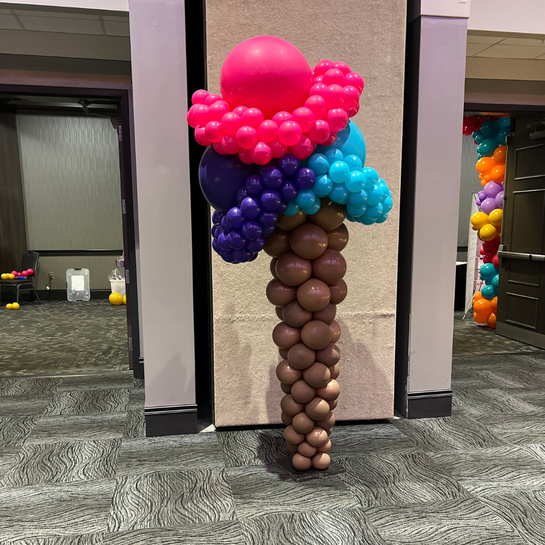 Colorful balloon sculpture resembling a large ice cream cone with a brown cone, blue ice cream, pink and purple toppings, and a large pink cherry on top, placed against a wall at an indoor event.