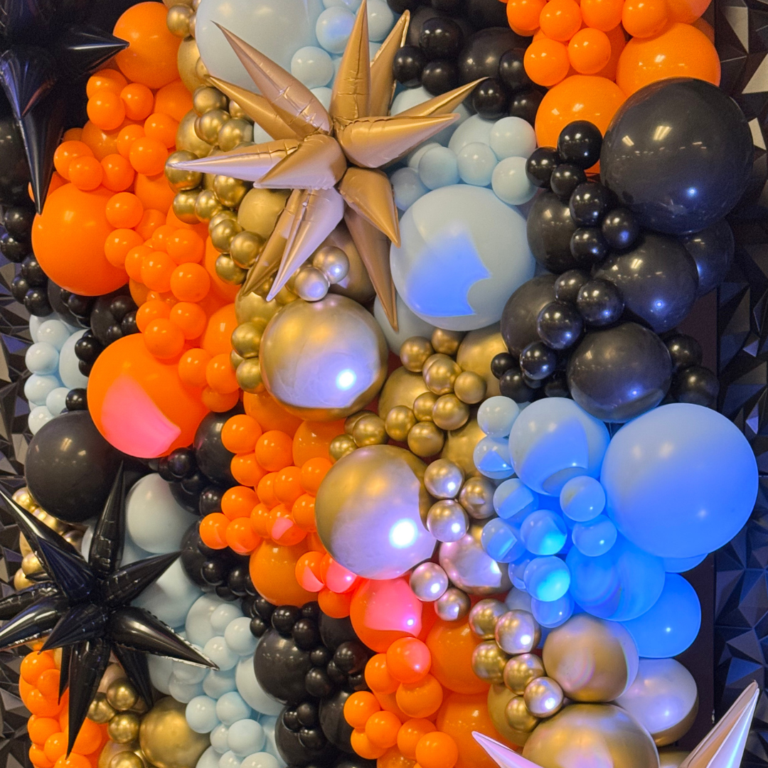 A colorful balloon arrangement with orange, black, white, and gold balloons, featuring various sizes and star-shaped balloons, creating a festive display.
