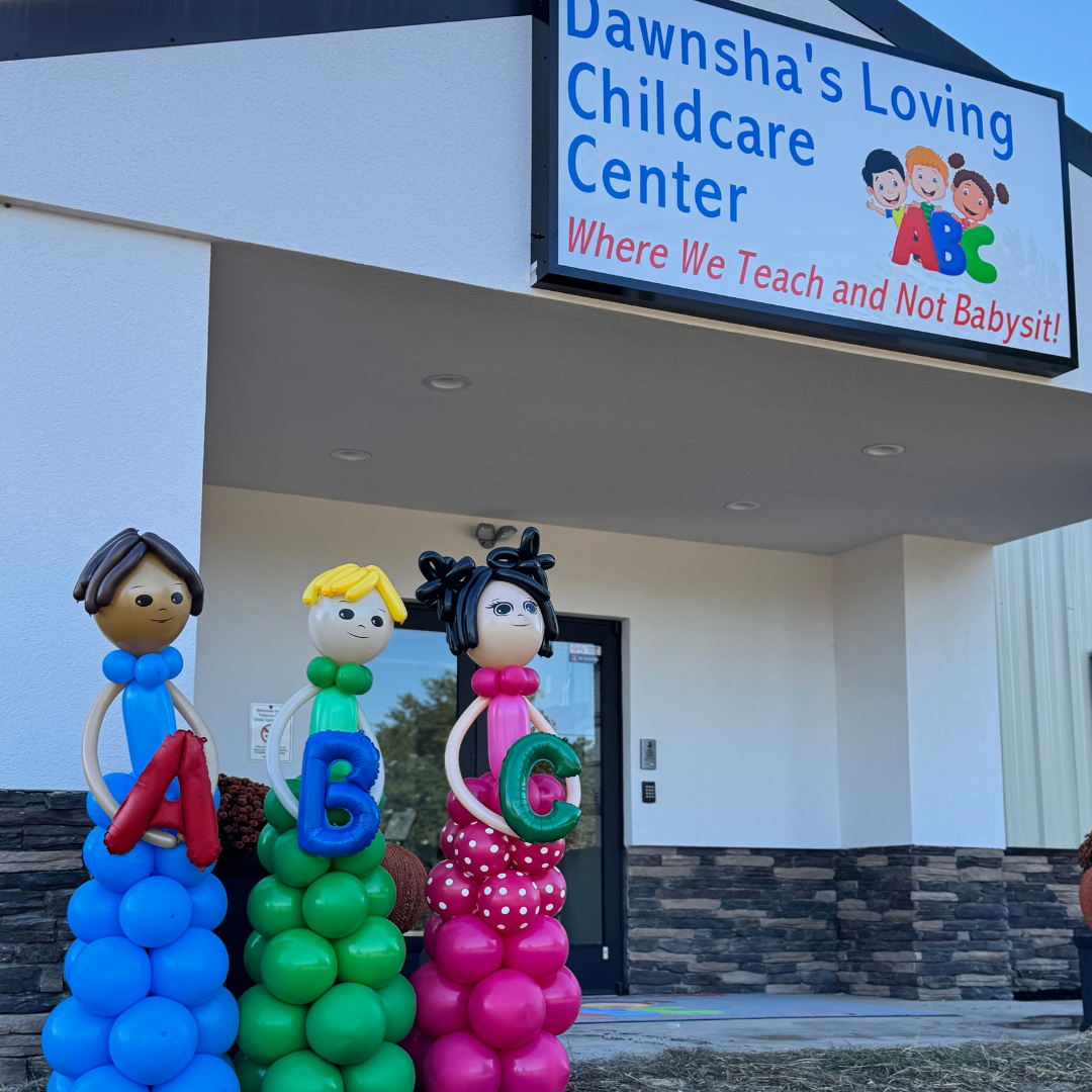 Colorful balloon sculptures of children holding letters A, B, C outside Dawnsha's Loving Childcare Center with a sign that says 'Where We Teach and Not Babysit!'