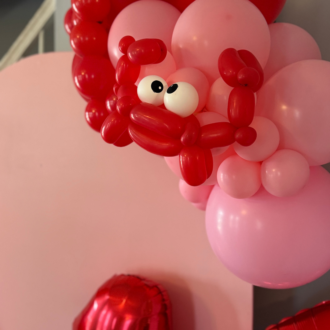 Balloon arrangement with pink and red balloons shaped into a lion's face, featuring large eyes and a mane of pink and red balloons, and a large pink balloon in the background.