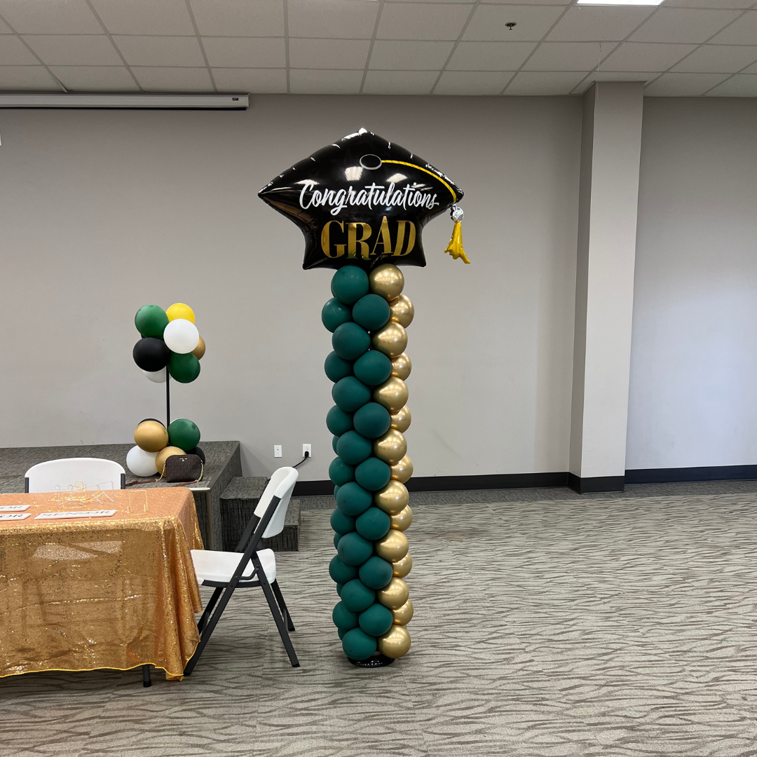 Balloon decoration with a graduation theme, featuring a column of green, gold, and black balloons topped with a black graduation cap-shaped balloon that has white and gold text saying "Congratulations Grad".
