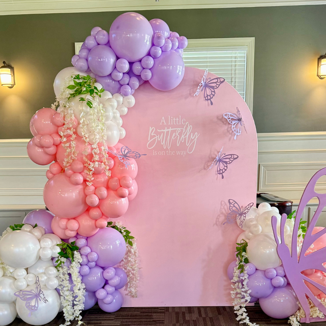 Decorative backdrop with pink, purple, and white balloons, white flowers, and butterfly cutouts. Text on the pink background reads "A little butterfly is on the way."