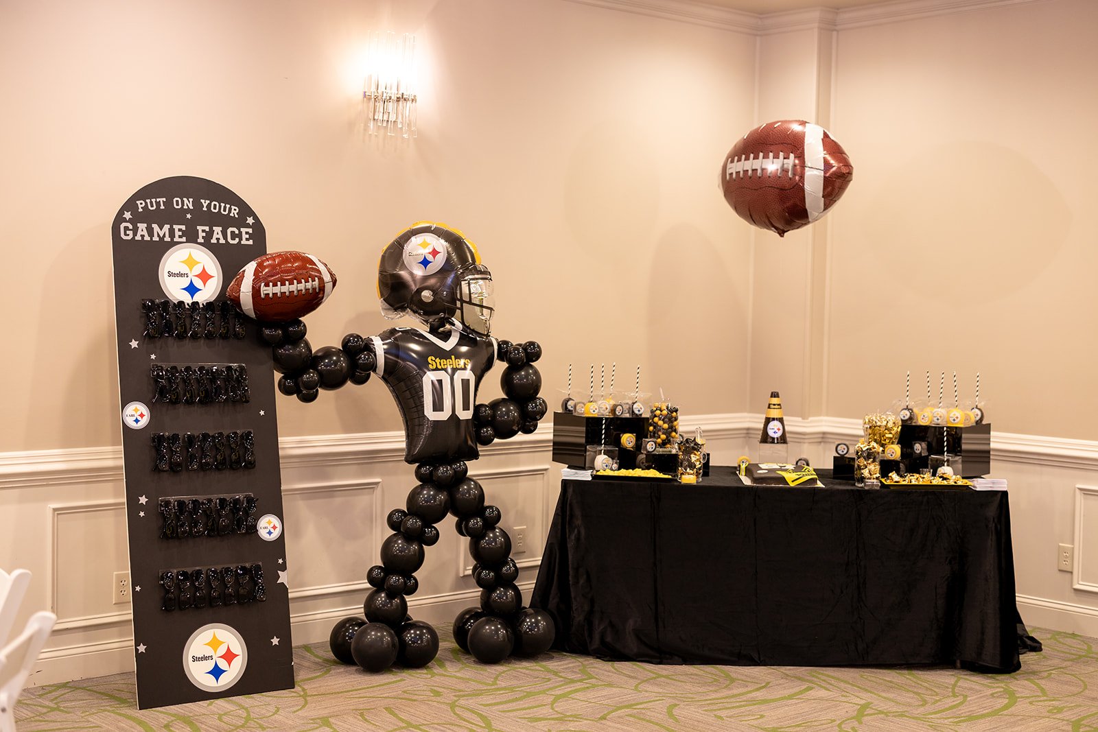 Party setup for a Pittsburgh Steelers themed celebration with black, yellow, and white decorations. Includes a balloon display, a balloon sculpture of a football player, and table decorations featuring Steelers logos and football-themed items.