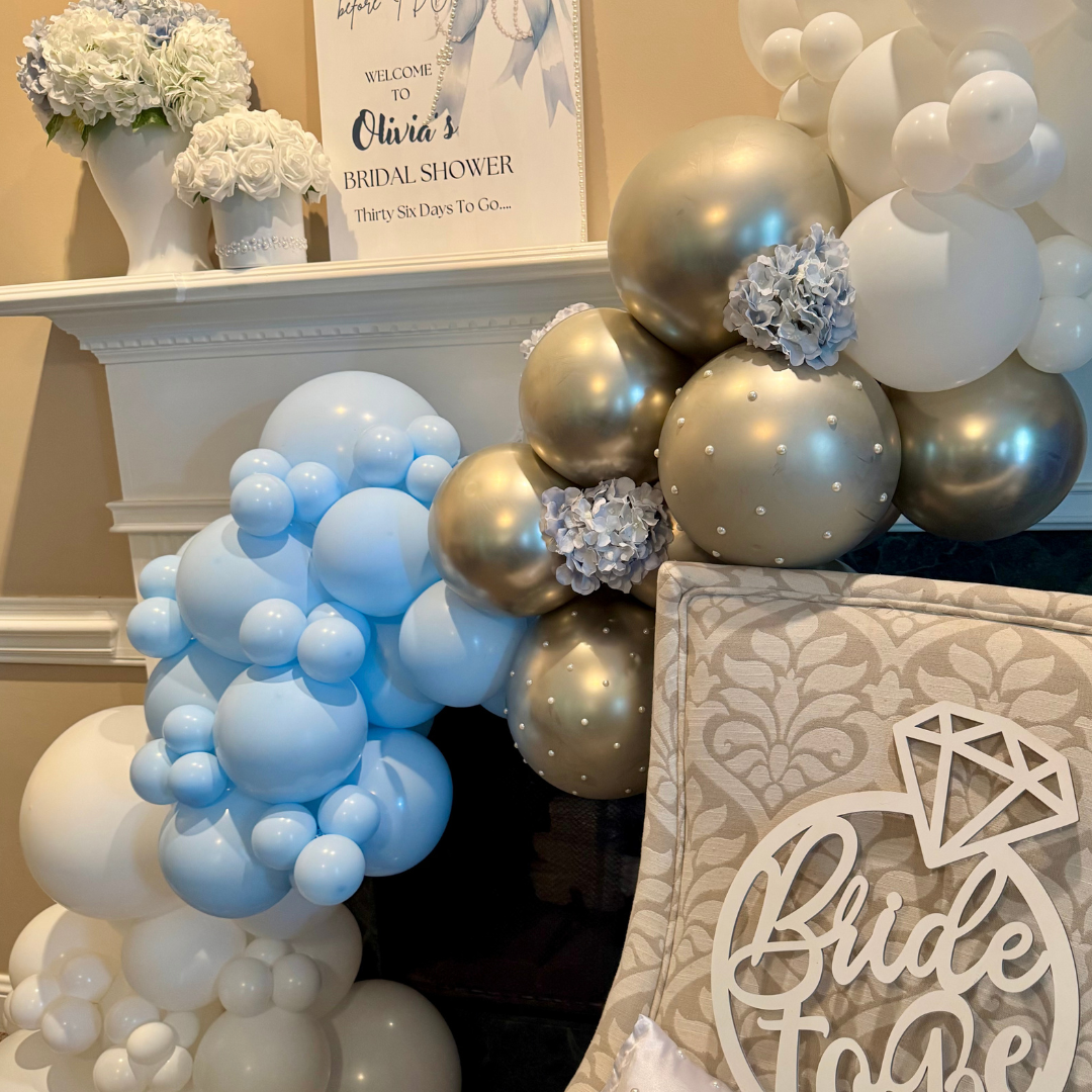 Decorative balloon arrangement in white, gold, and light blue for a bridal shower, with floral accents and a sign that reads "Welcome to Olivia's Bridal Shower" on the mantel, along with a "Bride to Be" sign.