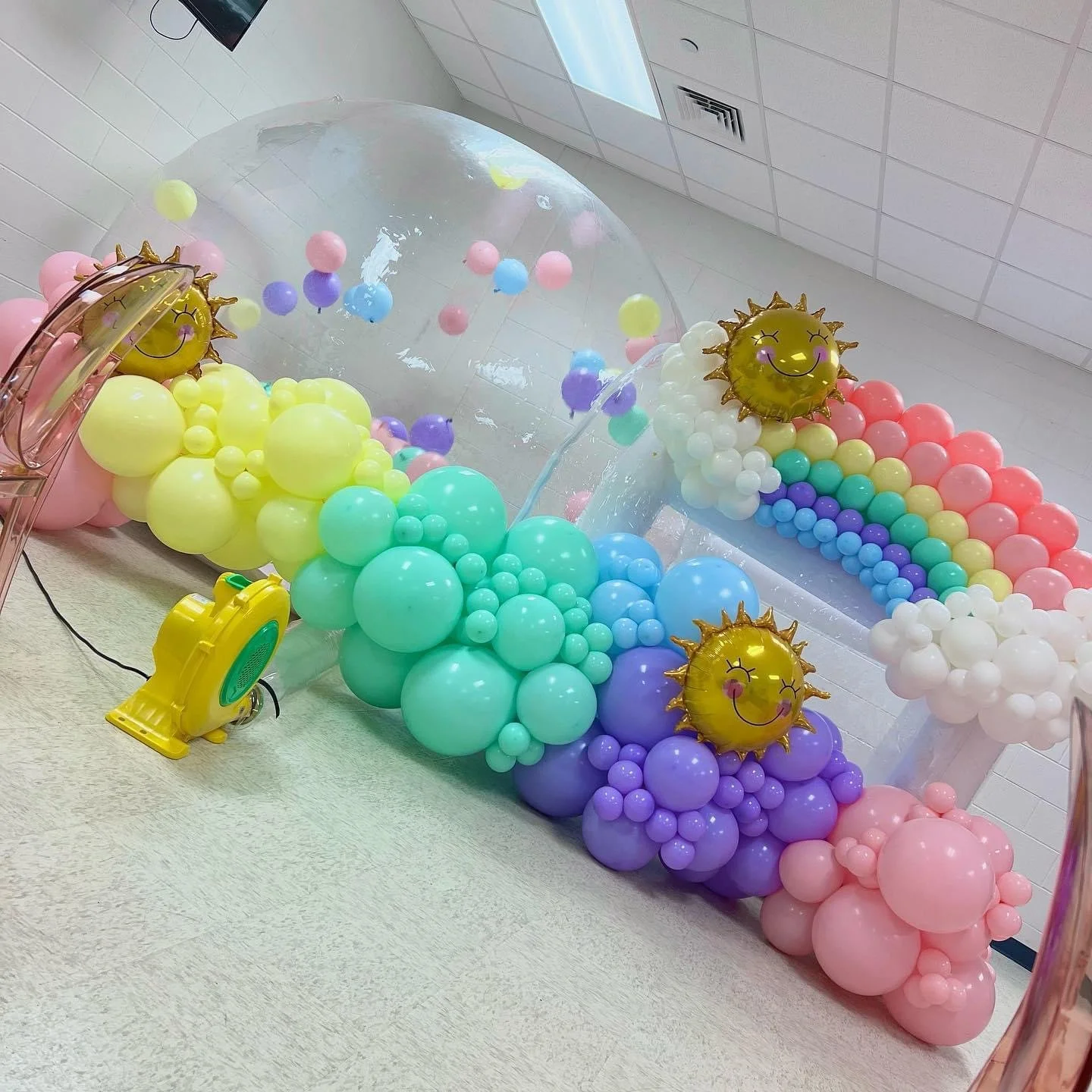 Colorful balloon arrangement with sun-shaped balloons, rainbow-themed balloons, and a transparent bubble arch situated on a floor in a room with white walls and ceiling.