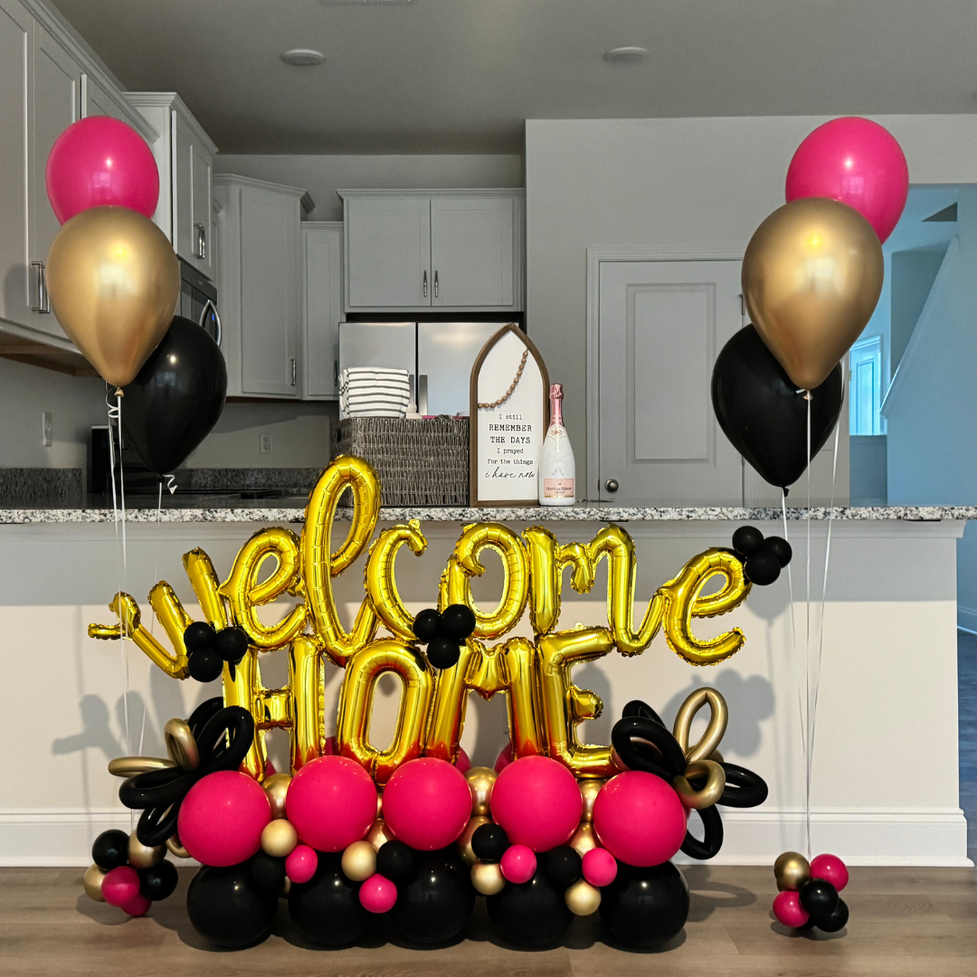 Decorative balloon display with pink, gold, and black balloons, including gold letter balloons spelling "welcome home," set up in a kitchen.