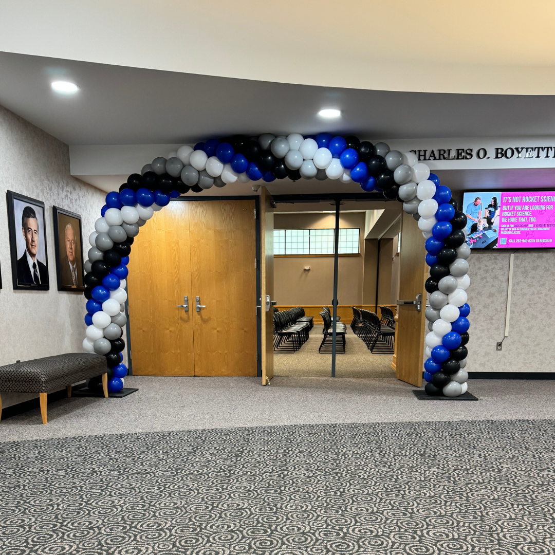 Decorative balloon arch in black, white, and blue at the entrance of a room with wooden doors, seating area, and portraits on the wall.