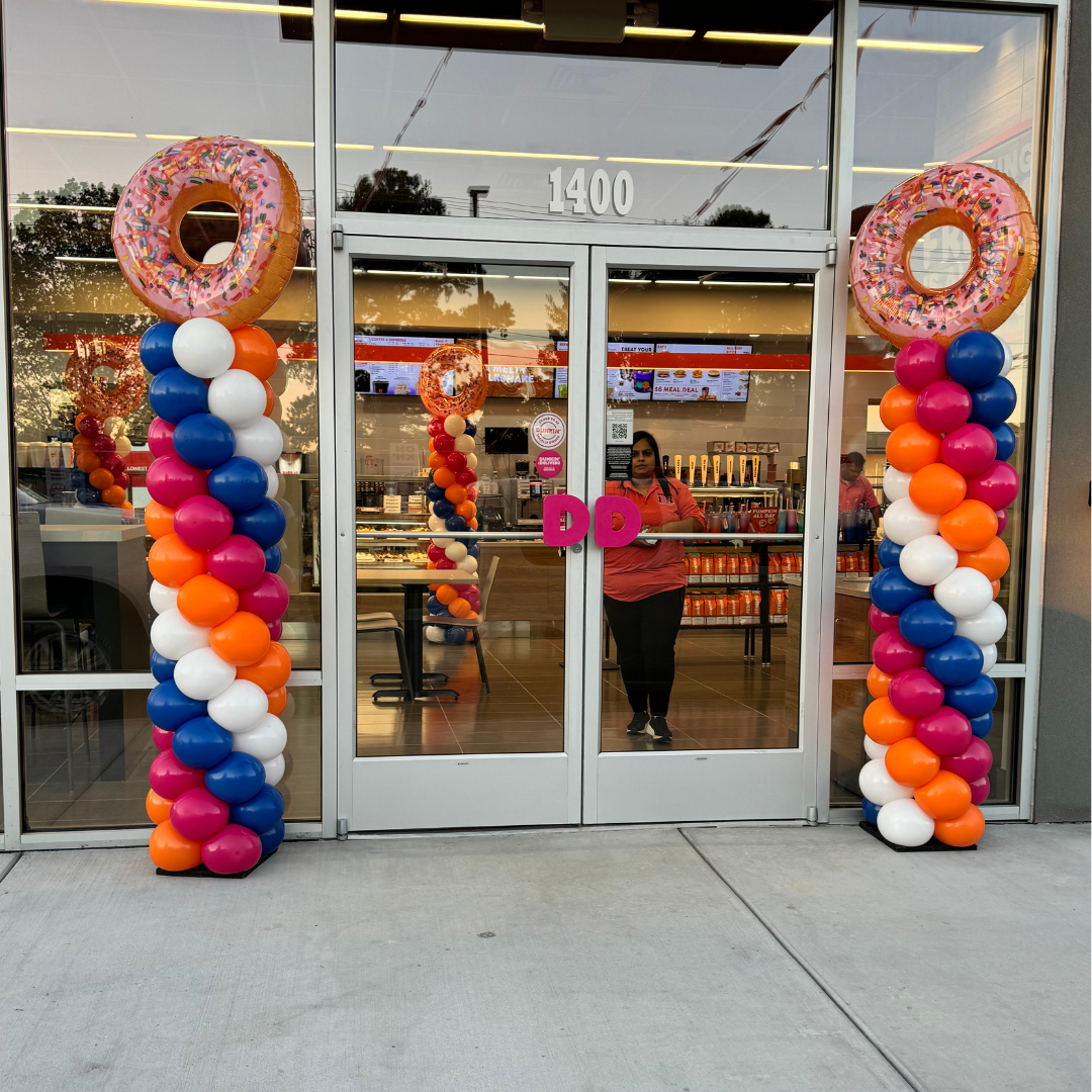 Colorful decoration outside a Dairy Queen store featuring two large balloon columns topped with donut-shaped balloons and a pink 'DD' sign on the glass door.