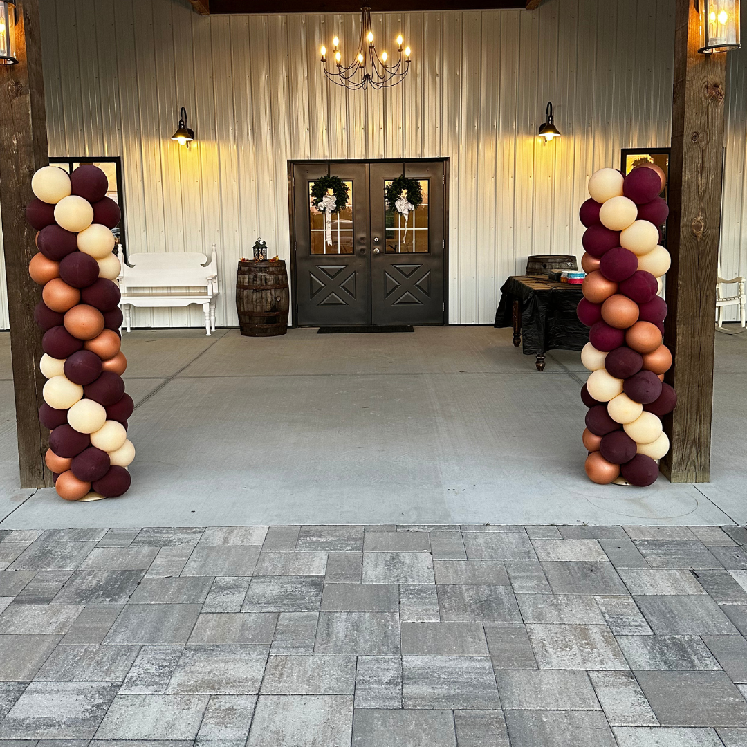 Decorated entrance with balloon columns on either side, featuring maroon, cream, blush, and copper balloons, leading to a set of black double doors adorned with white floral wreaths, in a rustic setting with industrial lighting.