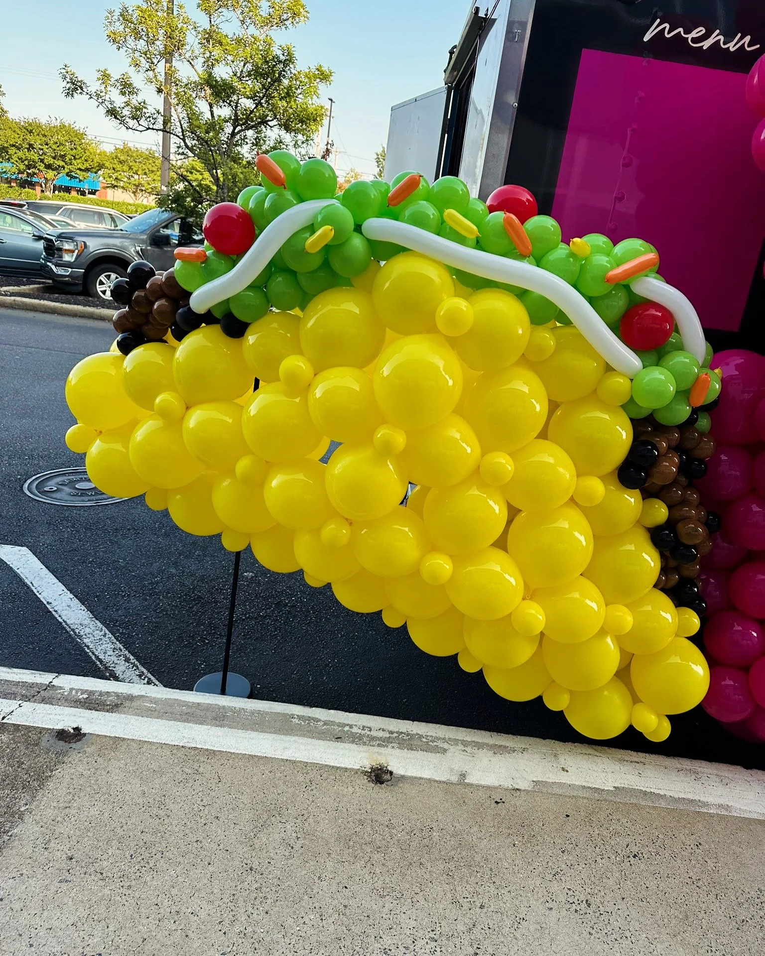 Balloon display resembling a bunch of bananas, with green, yellow, brown, red, pink, black, white, and orange balloons, outdoors in a parking lot.
