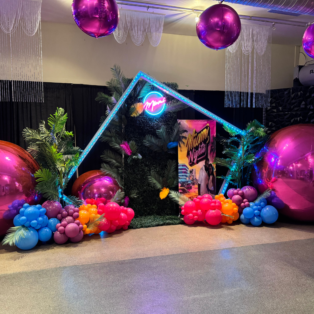 Decorative Miami Nights-themed backdrop with large pink and purple balloons, tropical plants, and neon signs, including a surfboard-shaped light with the Miami logo, for a festive event.