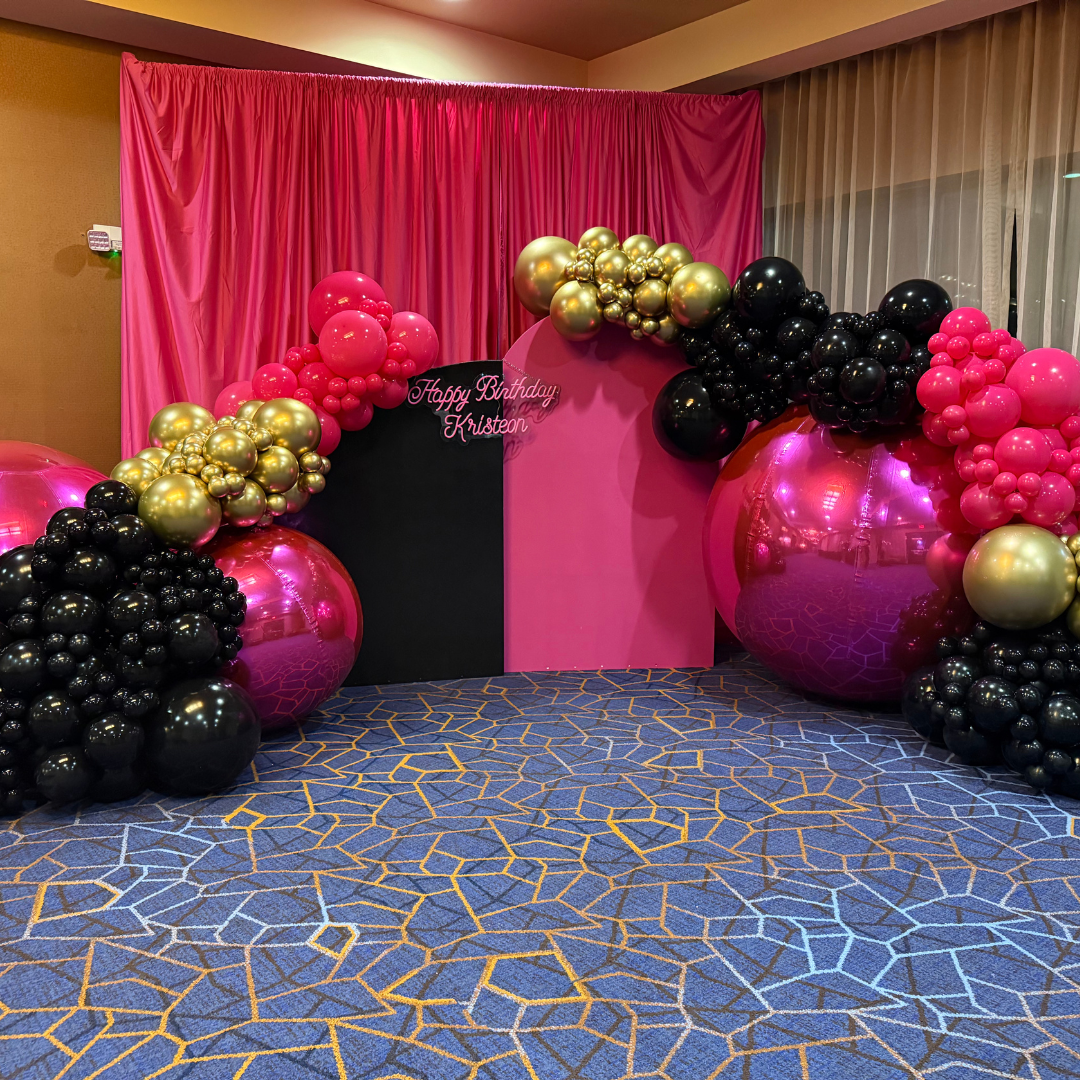 Balloon arch with pink, black, and gold balloons in front of a pink backdrop with a birthday message for Kriston.