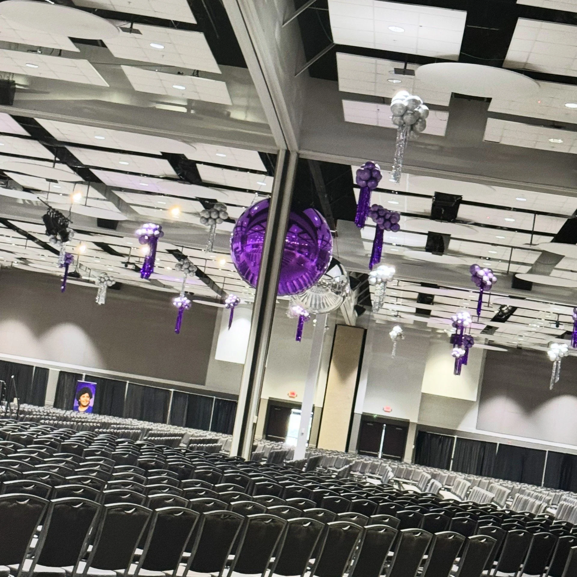 Empty banquet hall decorated with purple and silver balloons hanging from the ceiling, with rows of chairs set up for an event.