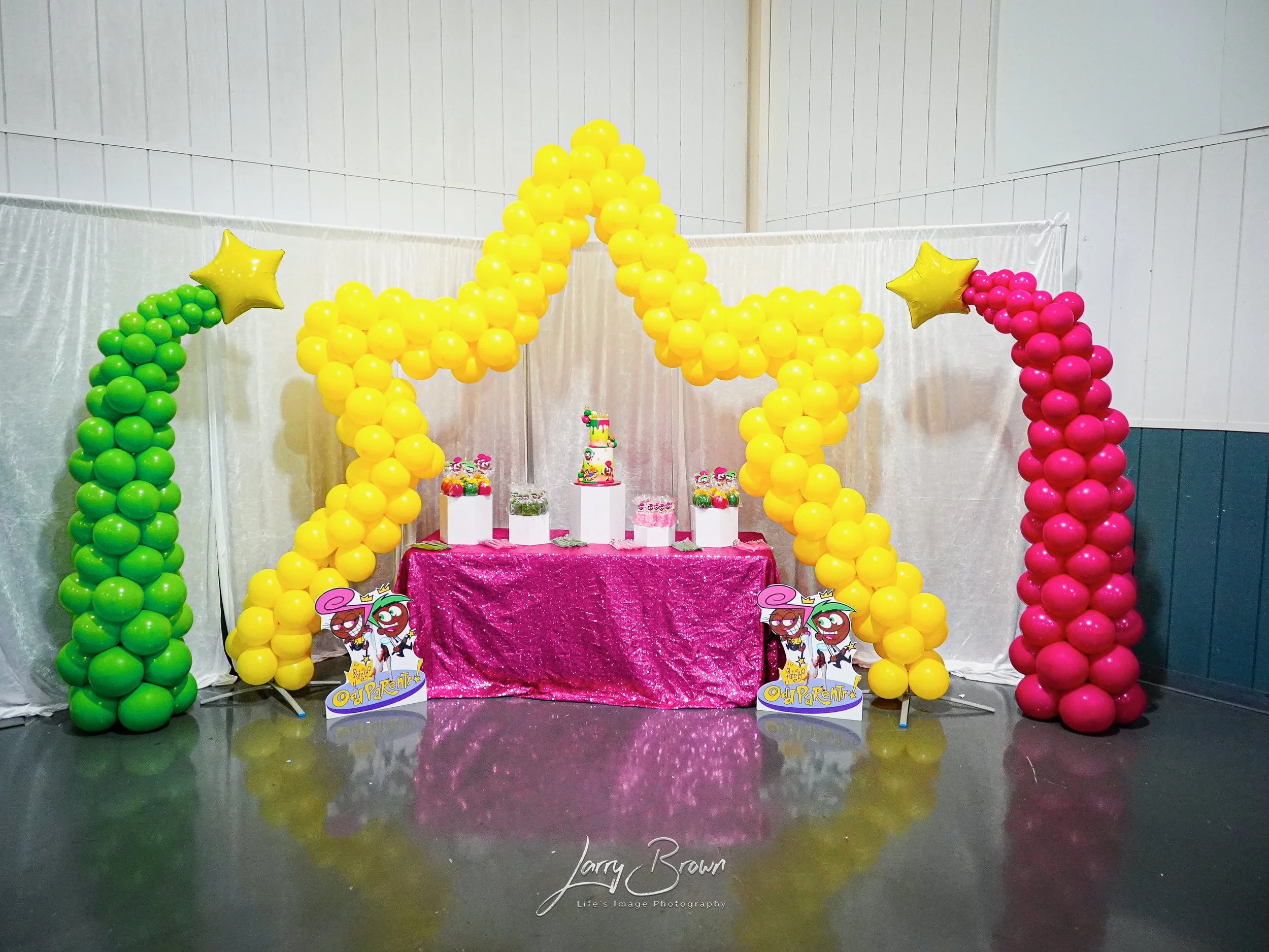Colorful balloon decoration with a yellow star-shaped balloon arch, pink and green balloon columns, a pink tablecloth, and decorated cakes for a birthday celebration.