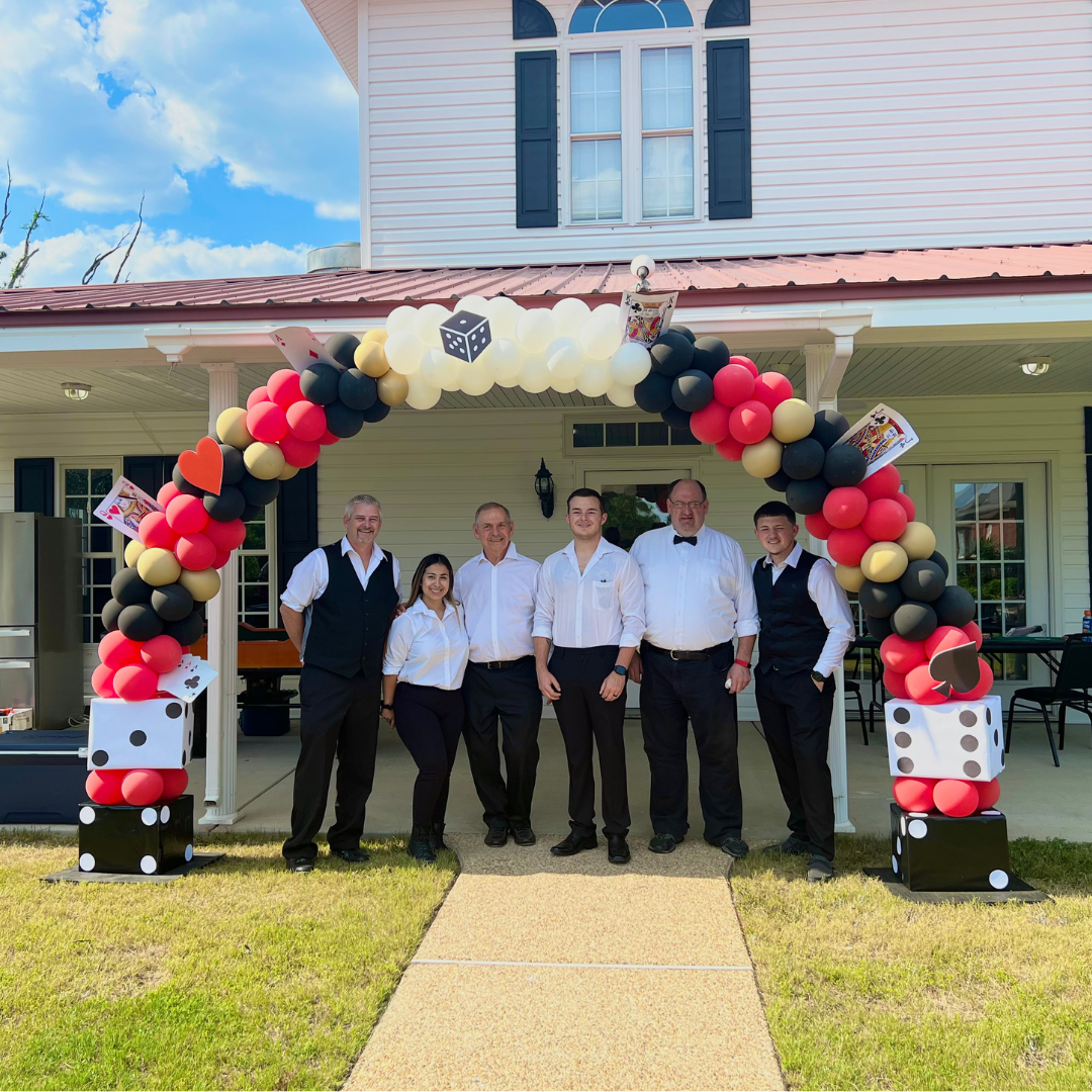 Group of six people dressed in formal attire standing under a decorative balloon arch with numbers, hearts, and die motifs, outside a house with a porch.