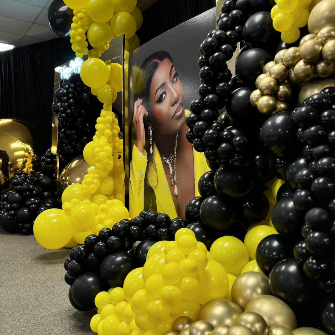 A large portrait of a woman wearing jewelry and a yellow dress, surrounded by black, yellow, and gold balloons at an event or celebration.
