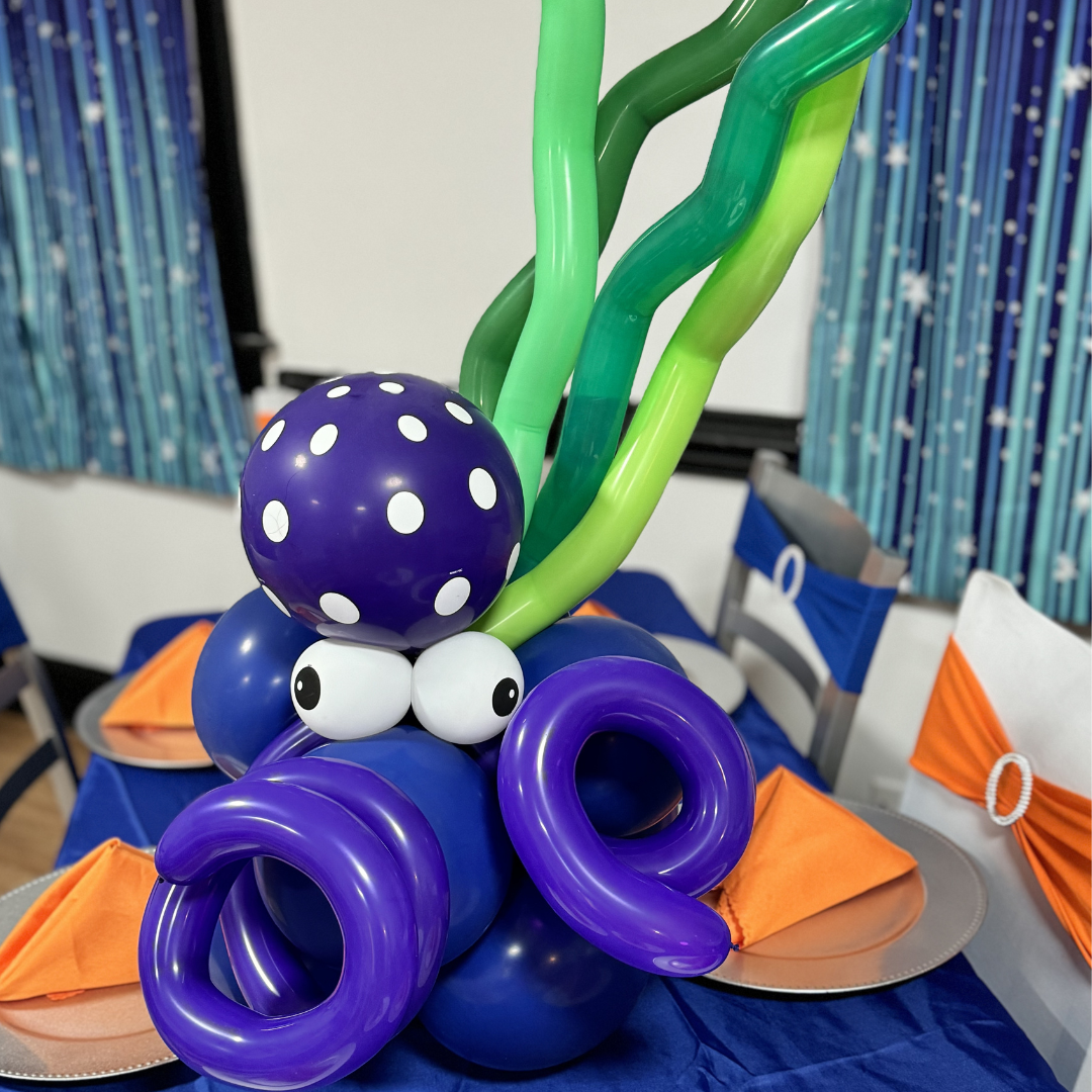 Balloon centerpiece featuring a purple octopus with green tentacles, purple and blue balloons, and a purple polka-dotted balloon head, set on a table with orange napkins and decorated chairs.