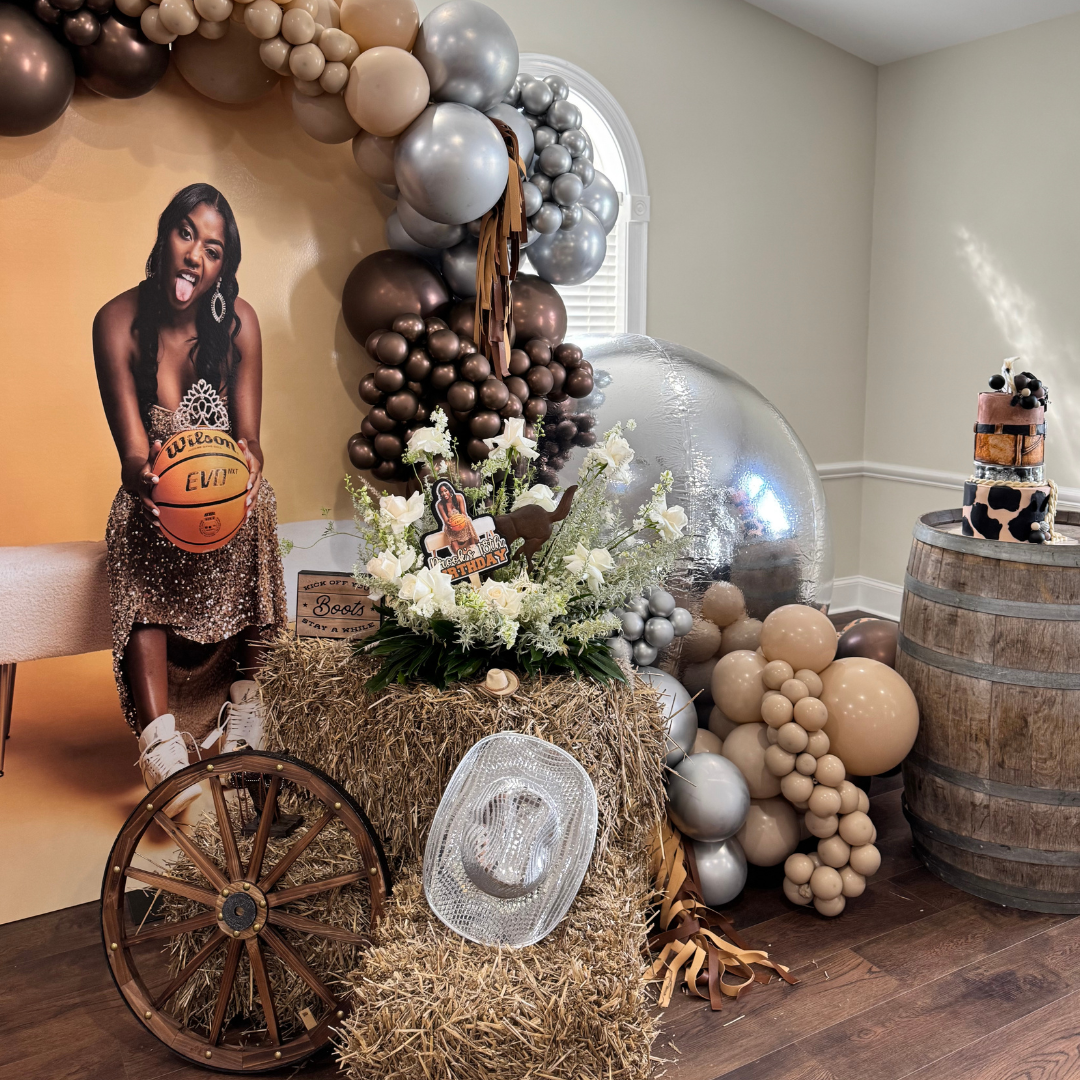 Decorative birthday party setup with a hay bale, white and brown balloons, a cardboard cutout of a girl holding a basketball, a floral arrangement, a transparent cowboy hat, and a cake on a barrel. The scene has farm-themed elements and a cow print d