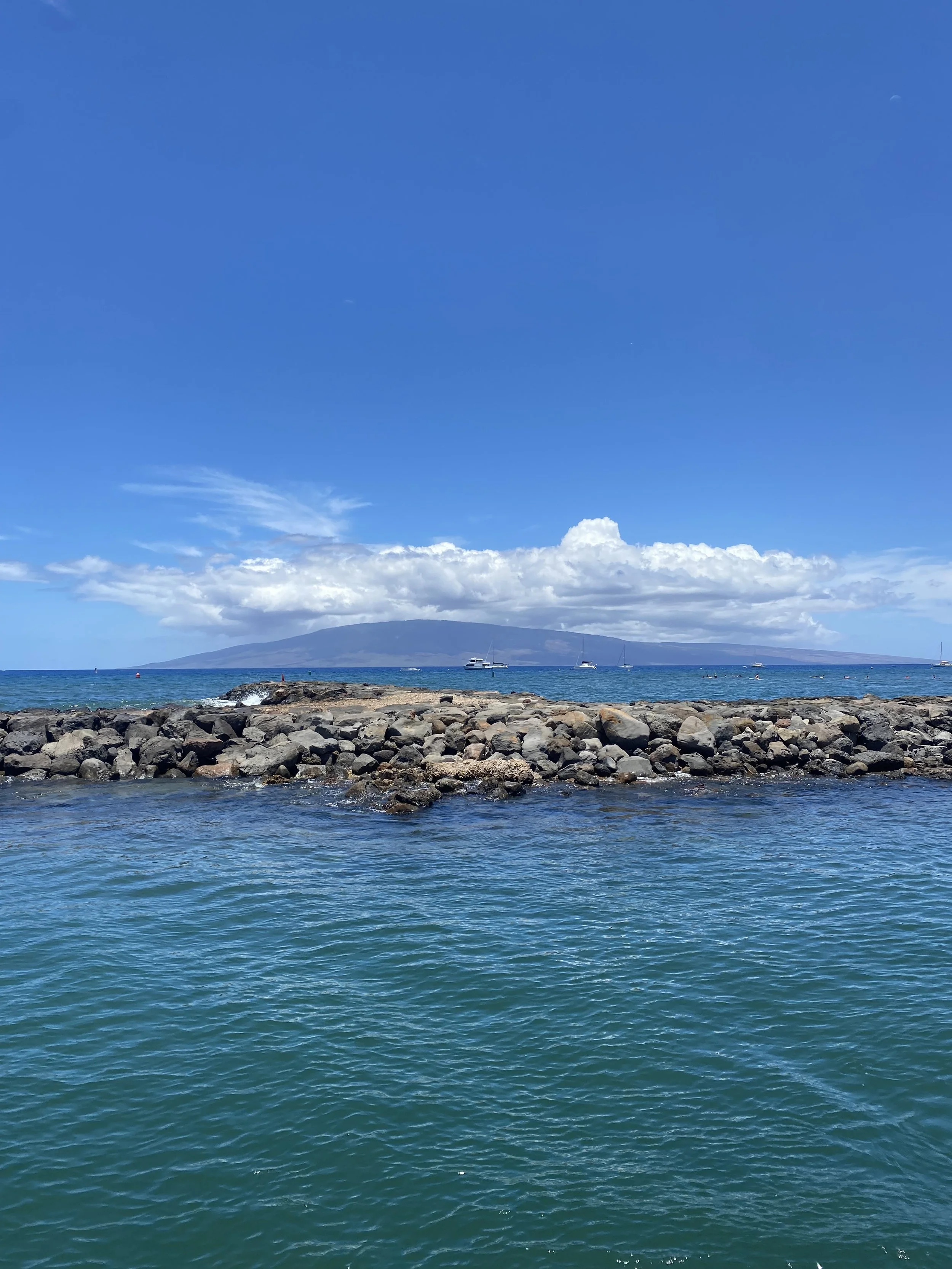 Atlantic Ocean with rocks forming a jetty, sailboats in the distance, large island or mountain in the background under a partly cloudy blue sky.