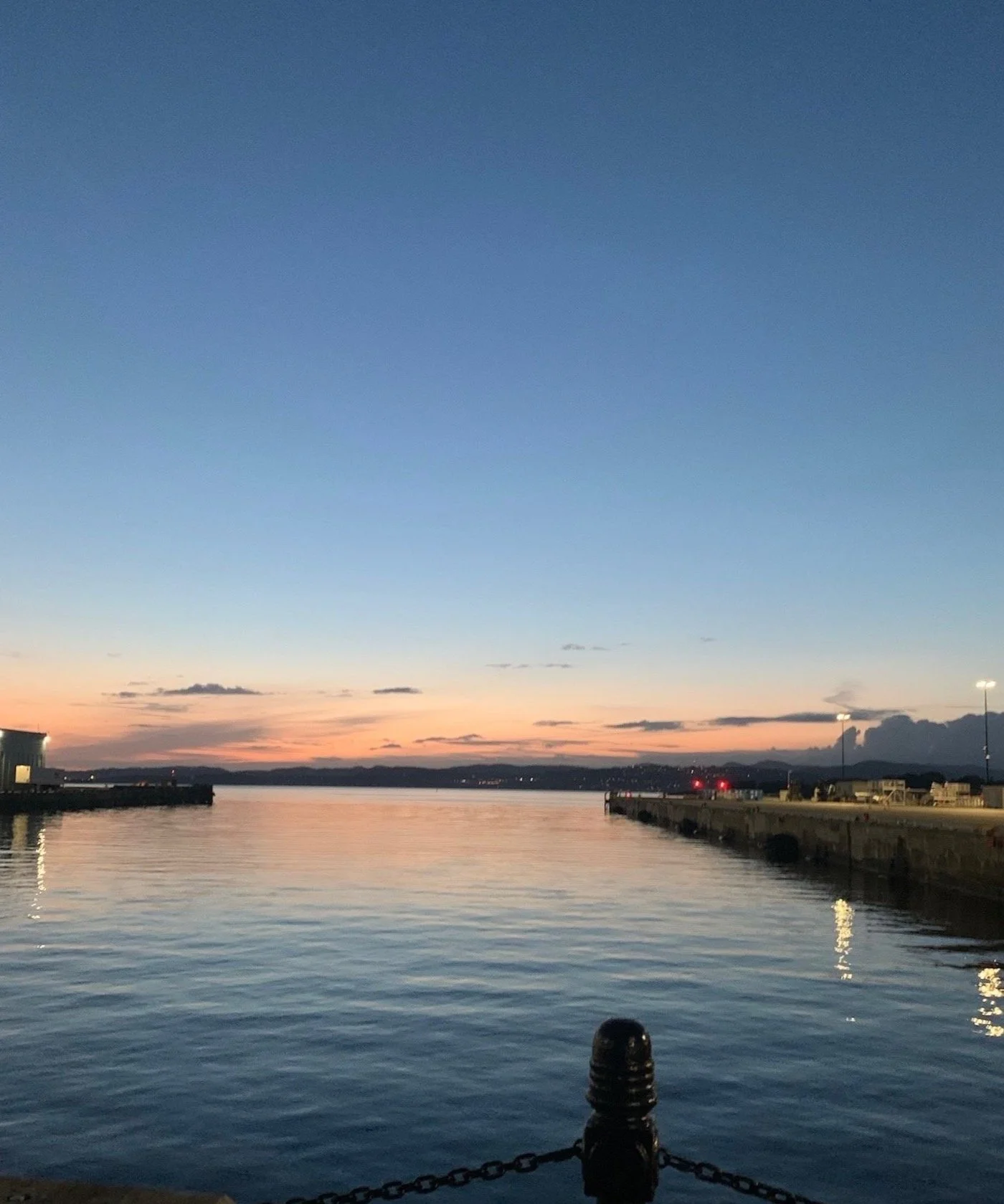 A serene harbor scene during sunset with calm water, distant hills, and a concrete pier on both sides, with lights on the pier illuminating the area.