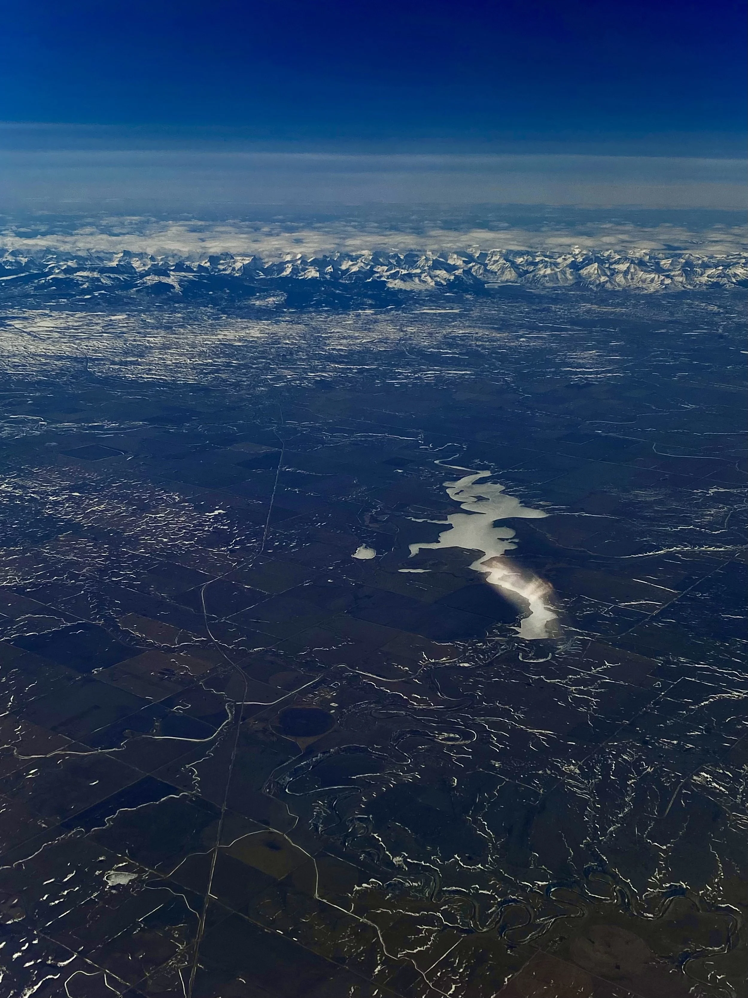 An aerial view of a landscape with a river, frozen lakes, and snow-capped mountains in the distance under a clear blue sky.