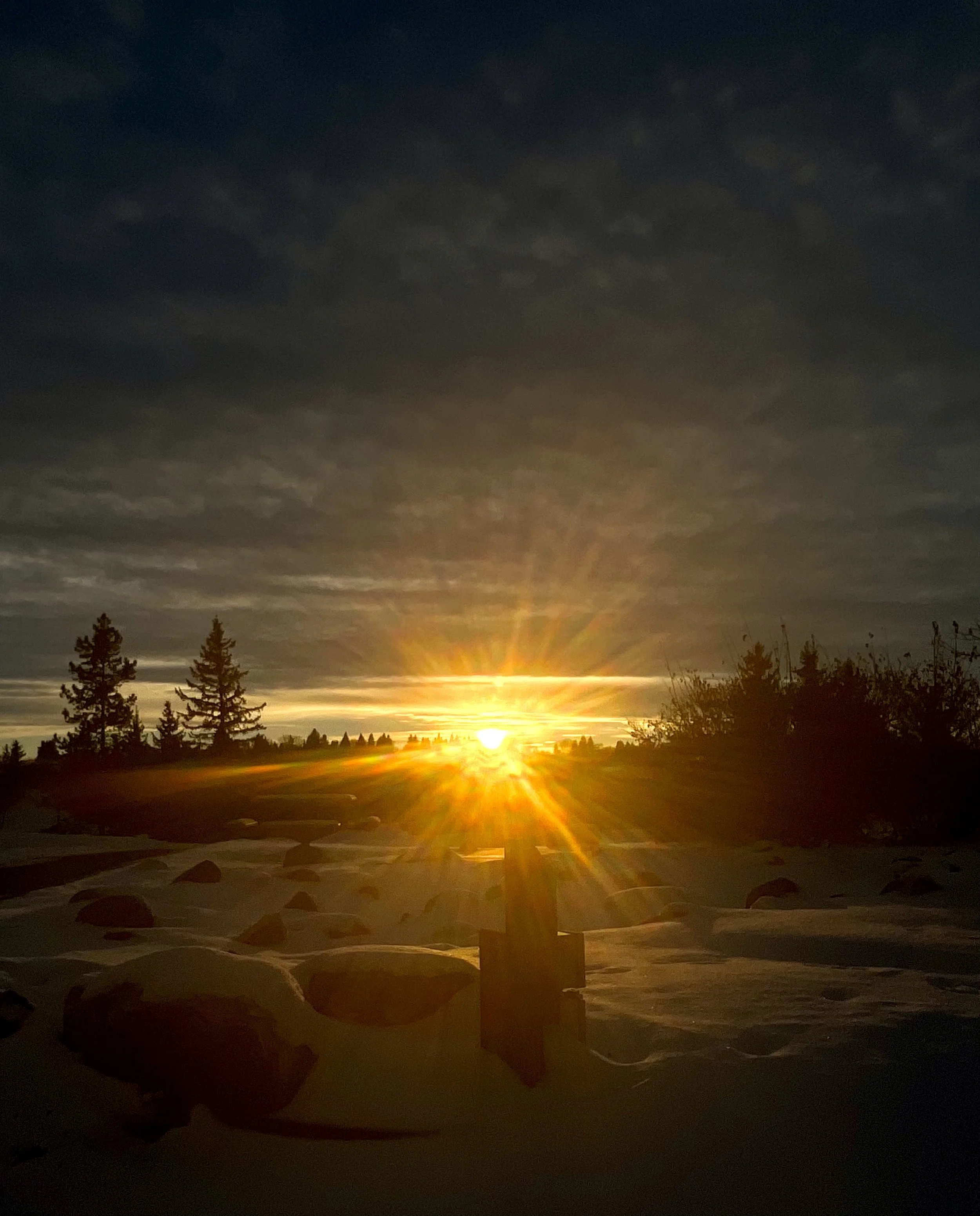 Sunset over a snow-covered landscape with trees and rocks, with an orange glow and sun rays stretching across the sky.