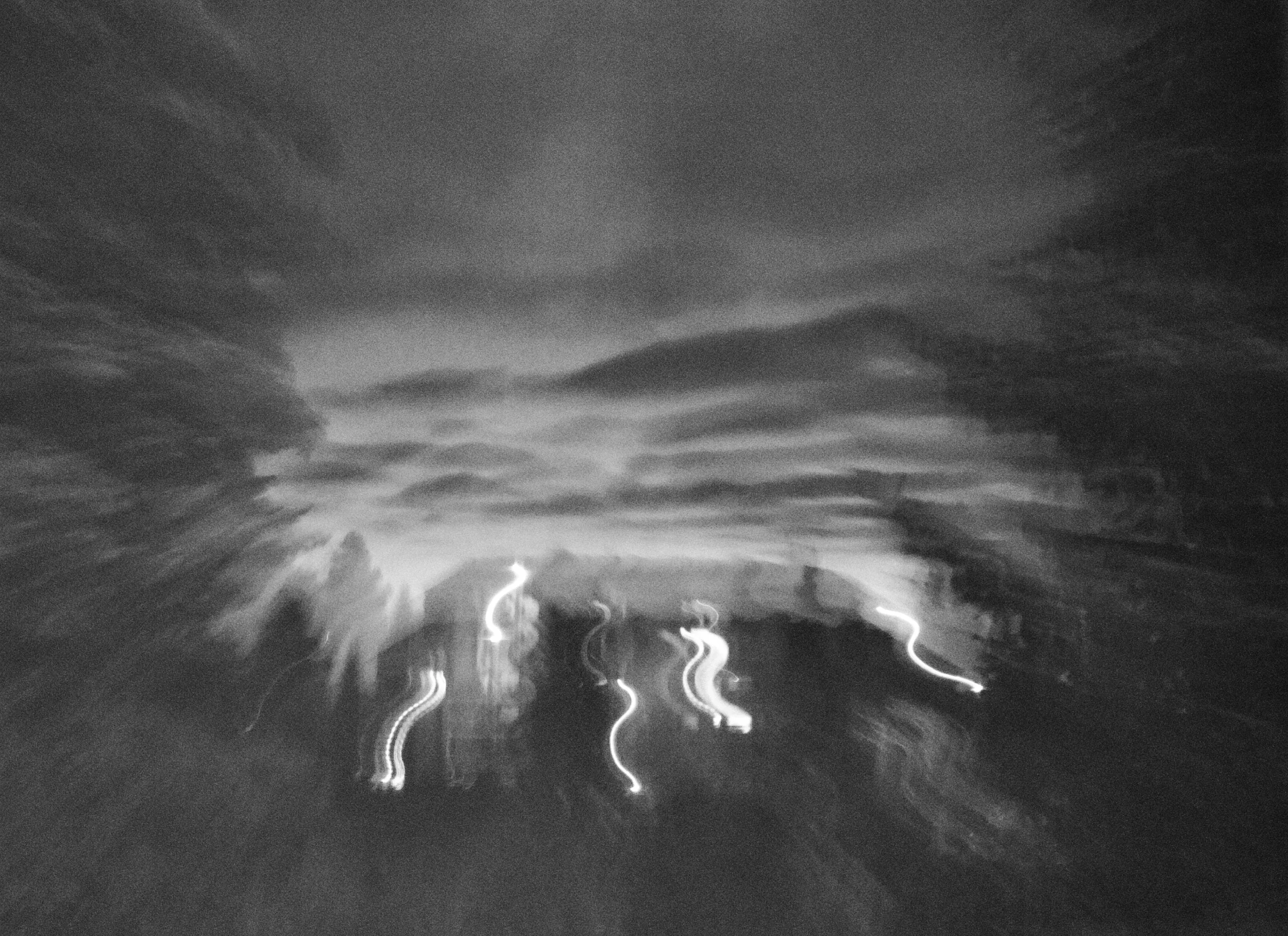 Blurry black and white image of a cloudy night sky with streaks of light, possibly from moving vehicles or streetlights.