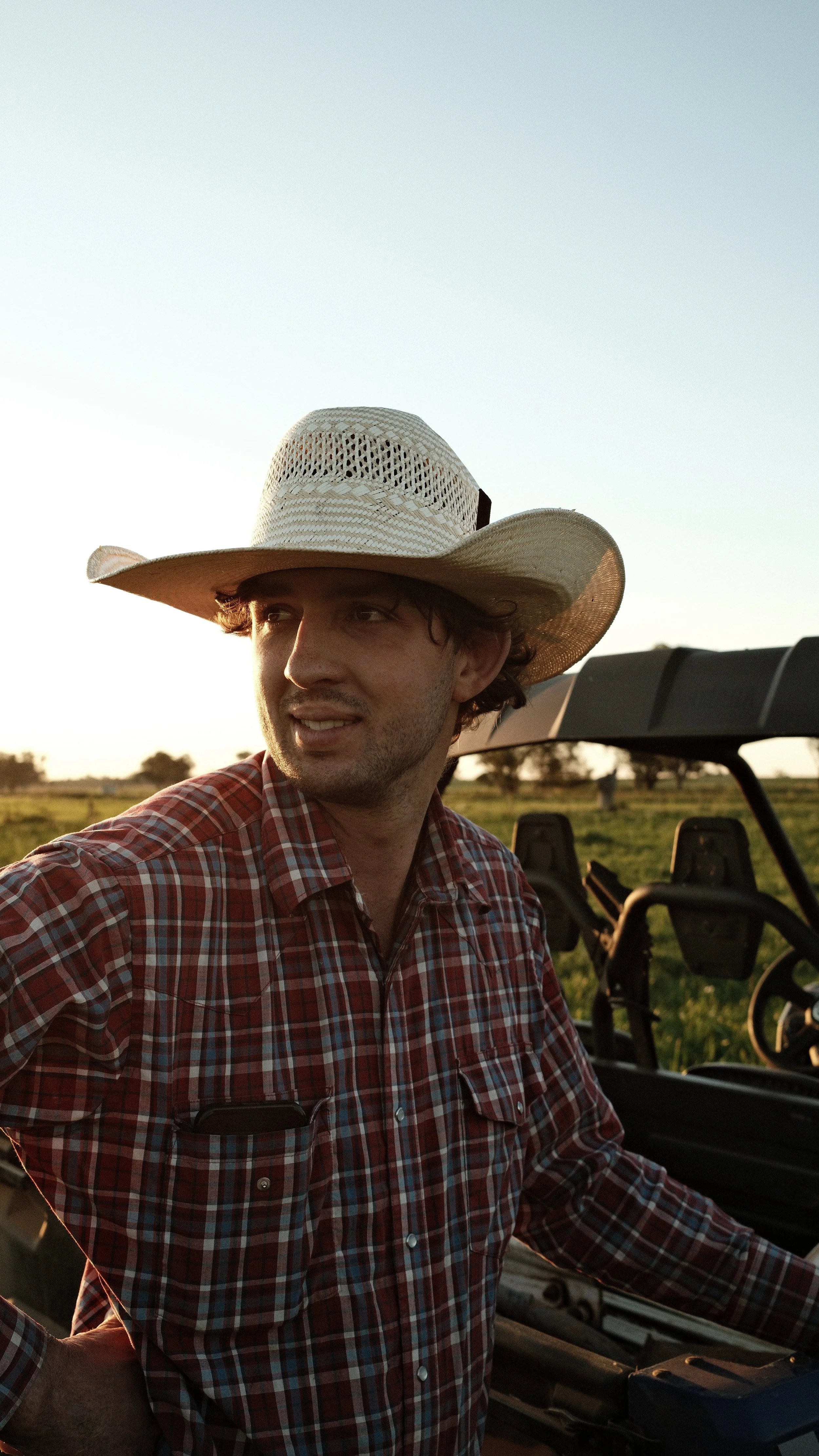 A man wearing a cowboy hat and plaid shirt standing in a field near a utility vehicle during sunset.