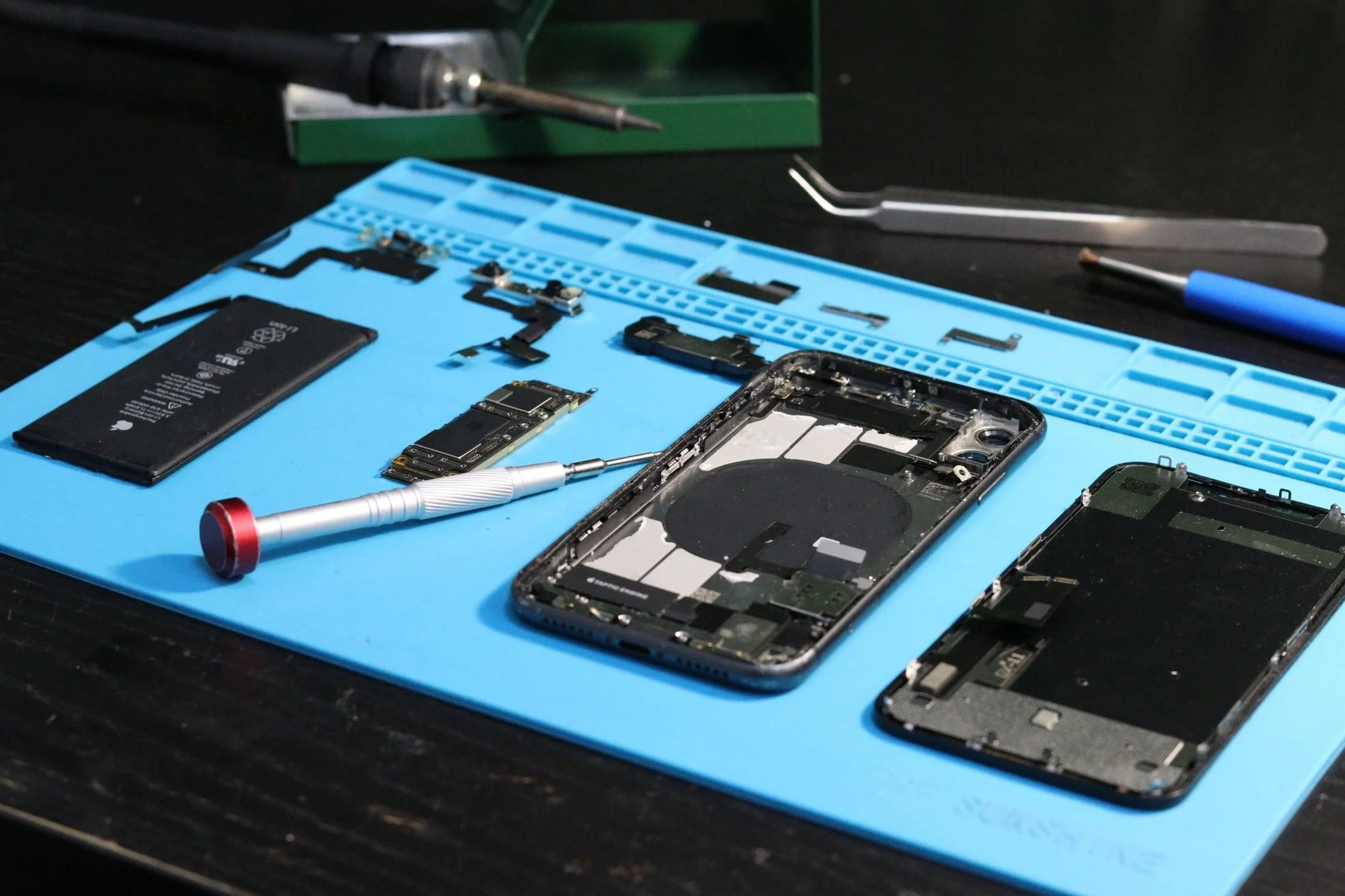 Disassembled smartphone on a blue anti-static mat with small electronic components, tools, and a soldering iron.