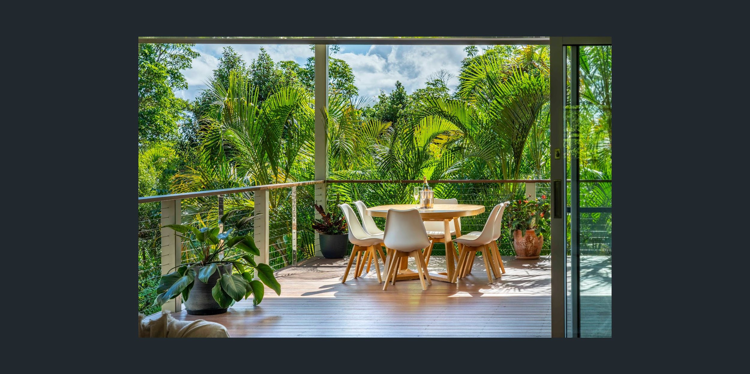 A cozy balcony with a round wooden table and six white chairs, decorated with potted plants, overlooking lush green tropical trees under a partly cloudy sky.