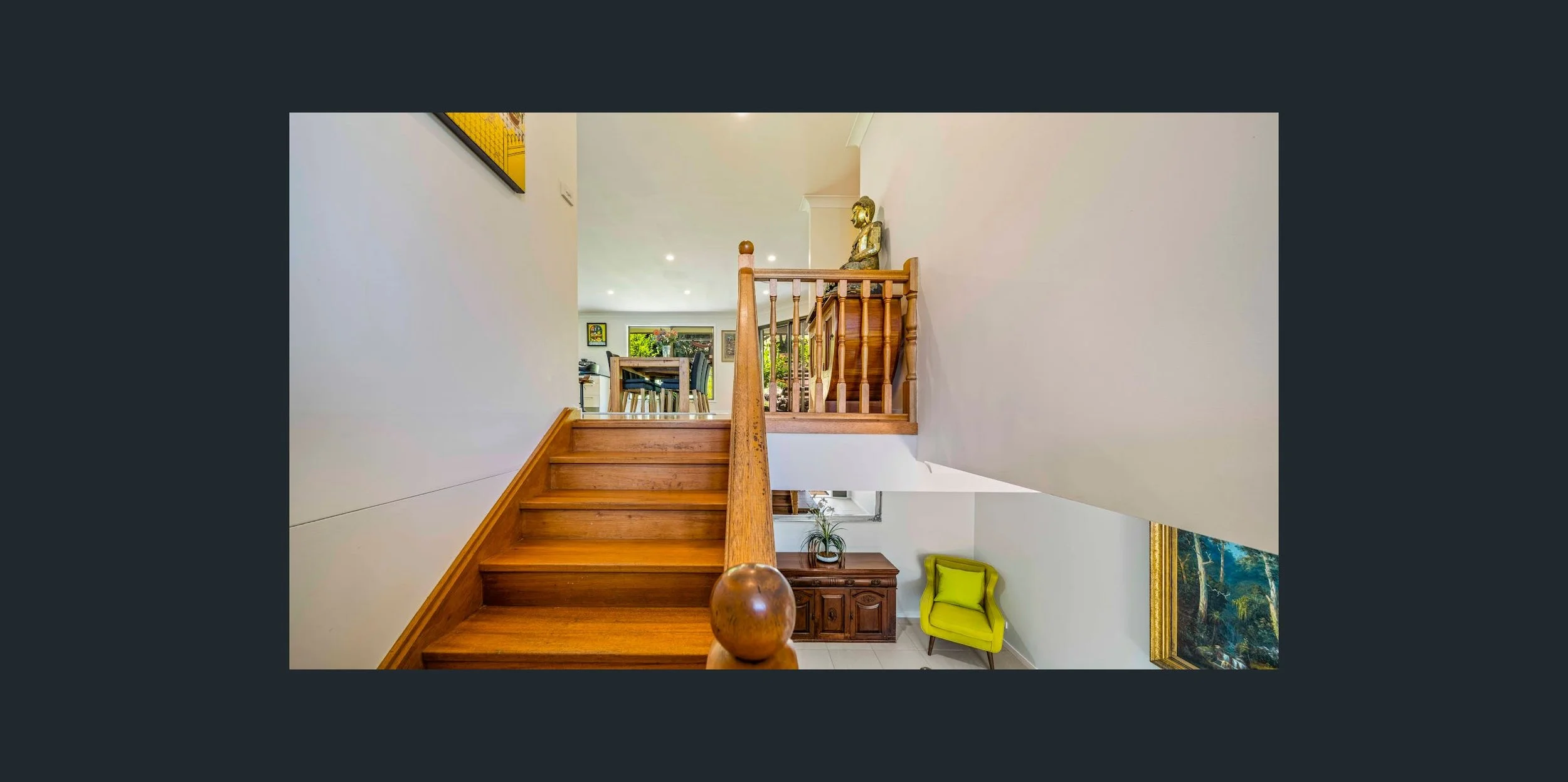 Interior view of a house staircase with wooden steps and railing, leading to a second floor. The first floor has a yellow armchair, a wooden cabinet, and a landscape painting on the wall. The second floor has a dining area with a table and chairs, an
